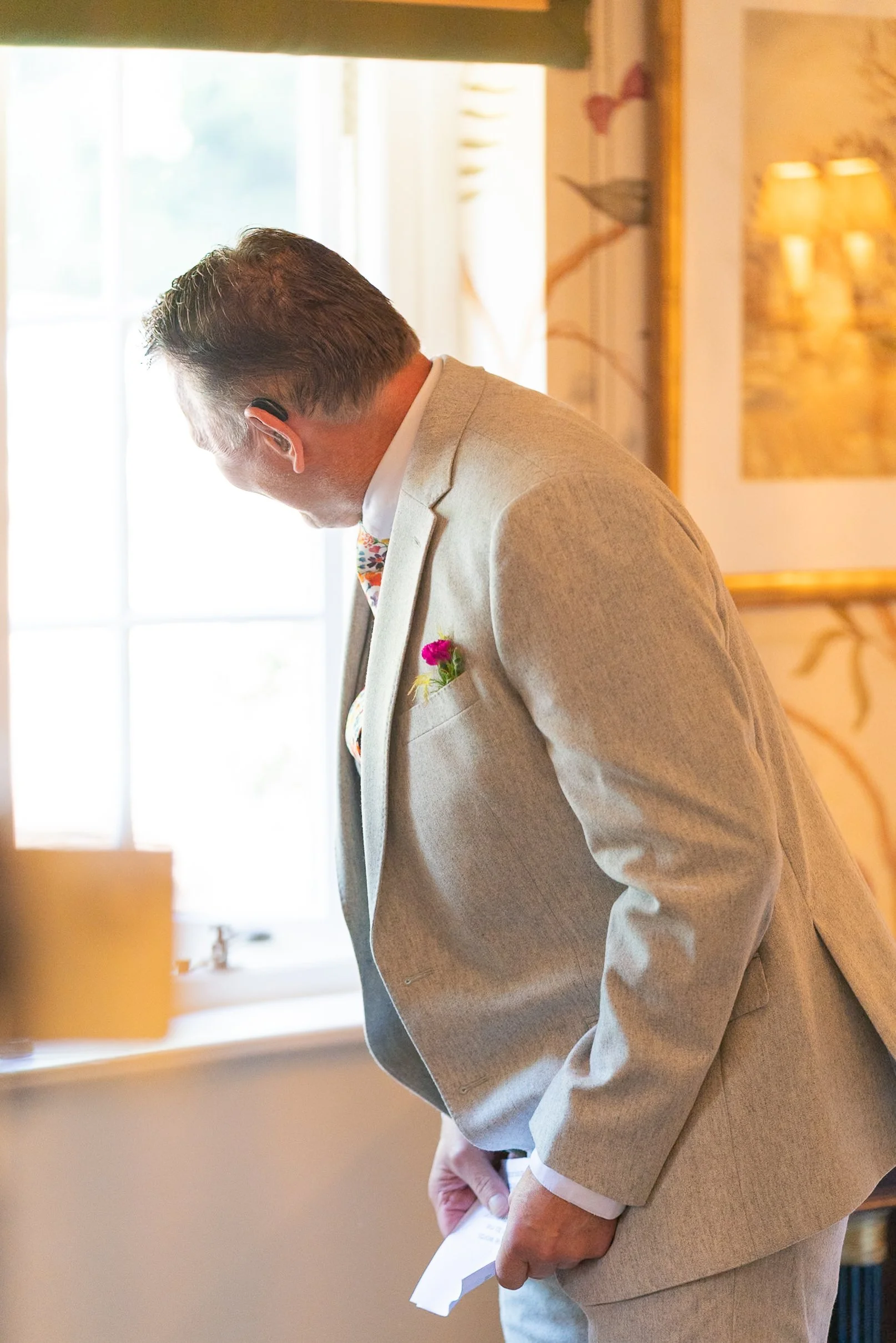 A man wearing a light-colored suit with a pink boutonniere is bowing inside a well-lit room with decorative wall art and a window.