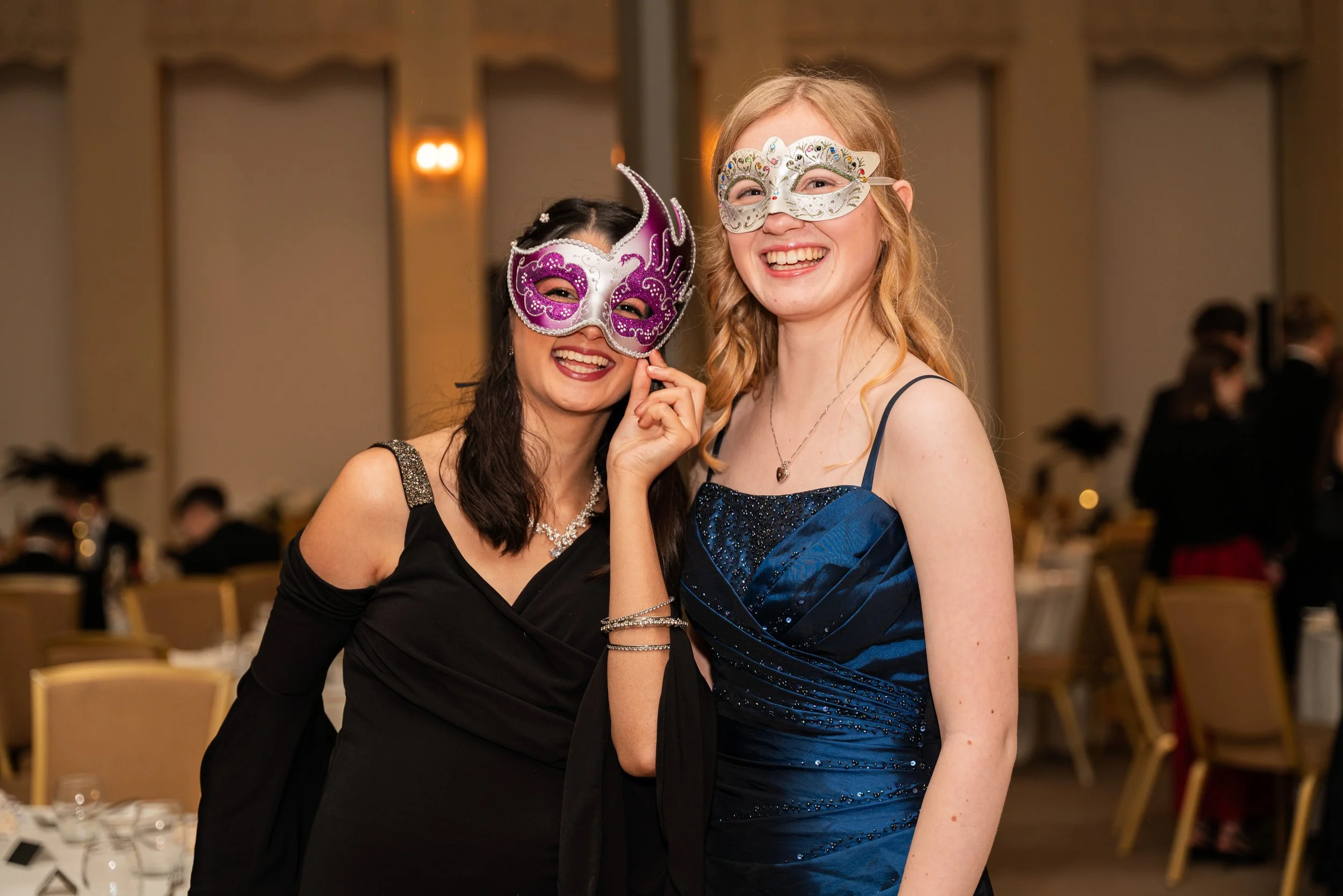 Two women wearing elegant dresses and masquerade masks smiling at a celebration or party.