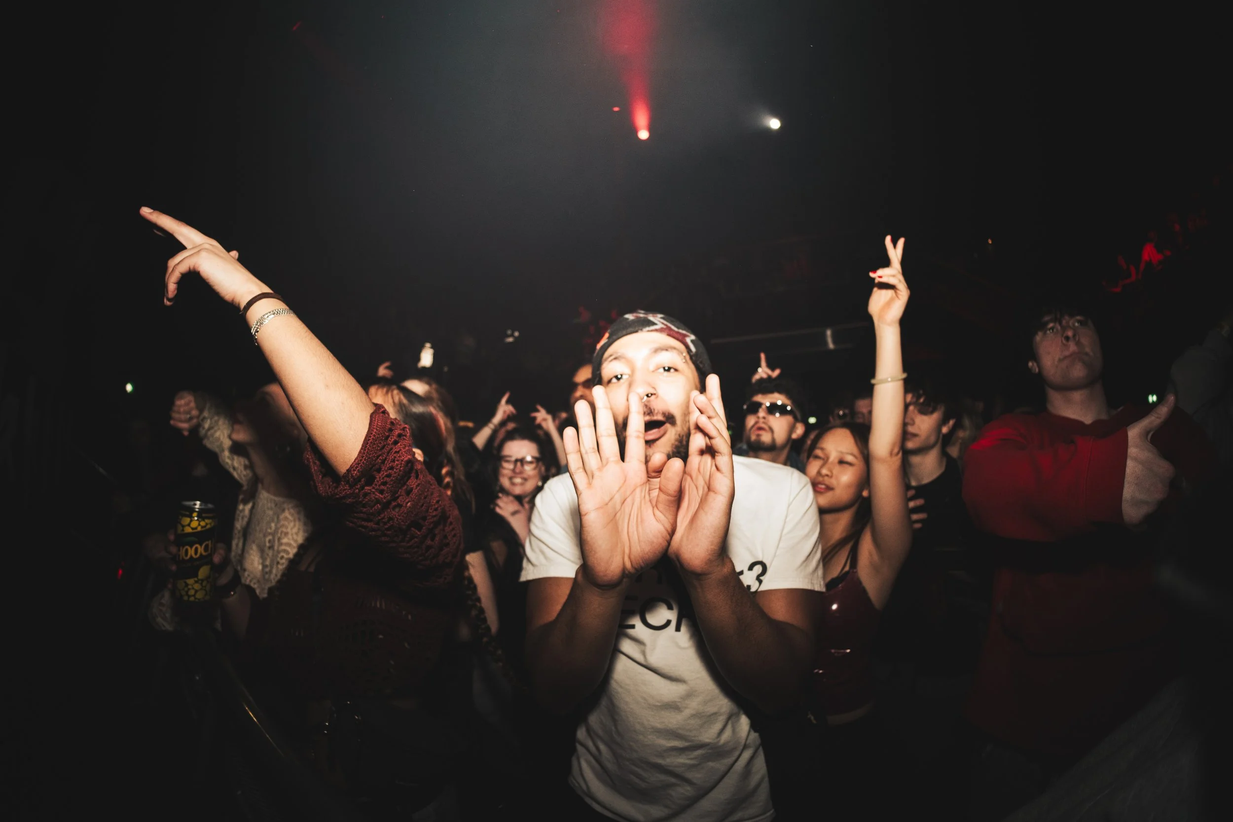 Crowd of people at a concert or nightclub, with one man in the center making a playful gesture with his hands near his face, others raising their hands or giving thumbs up, dark lighting, and red stage lights in the background.