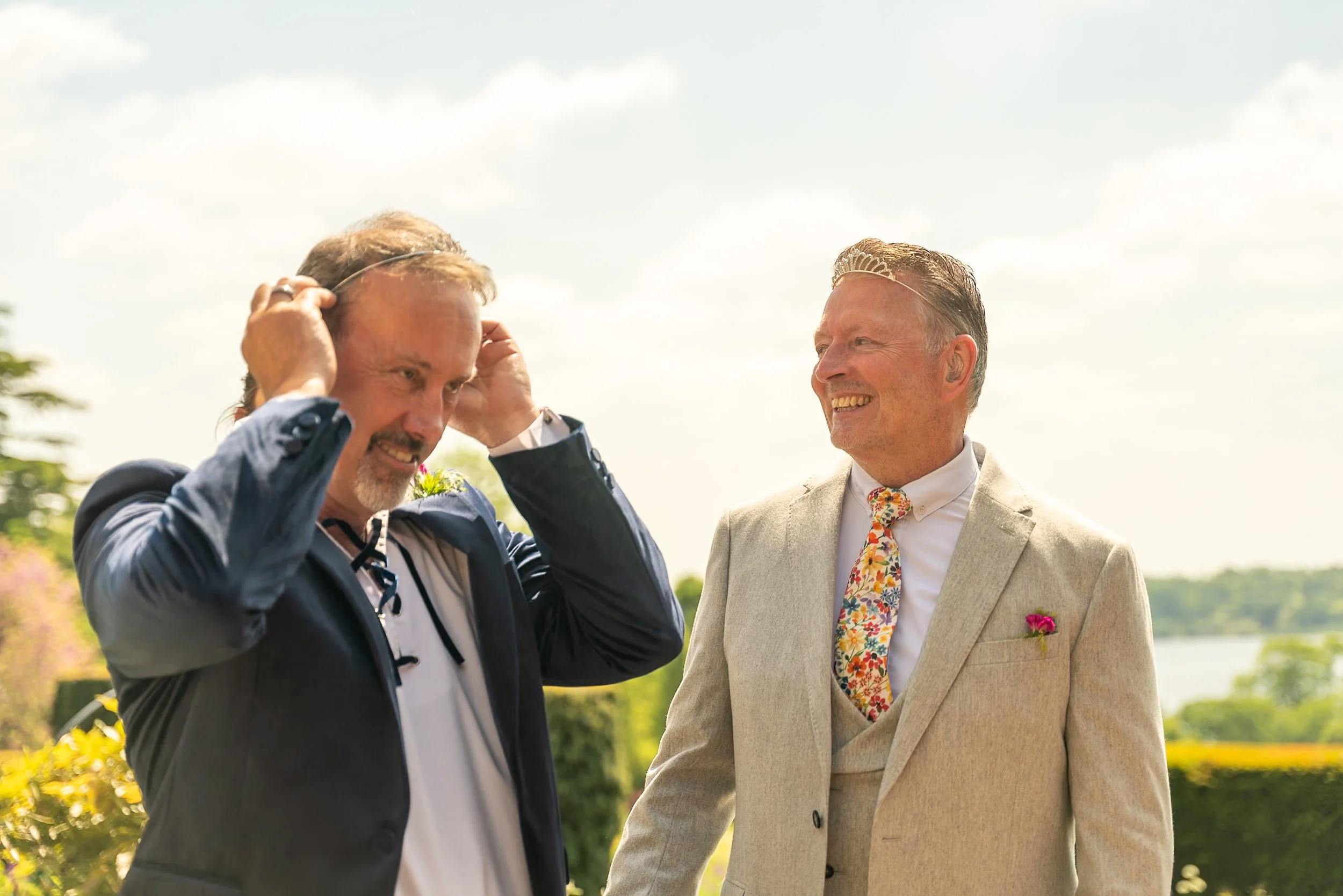 Two men dressed in formal attire are outdoors, smiling and interacting, with a scenic background of greenery and water.