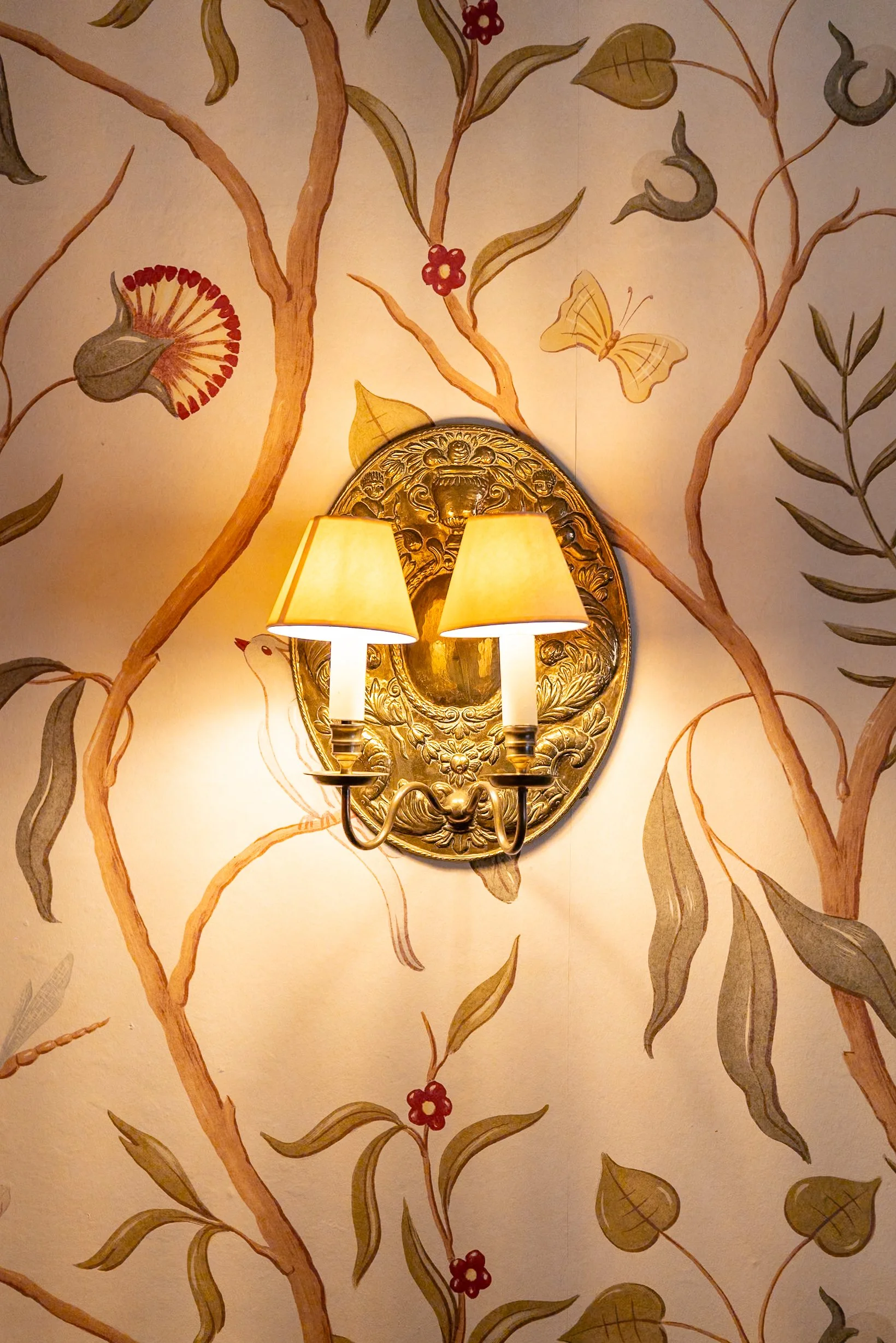 Wall with floral wallpaper featuring branches, leaves, red flowers, a butterfly, and a small bird. A gold ornate wall sconce with two lamps is mounted in the center.