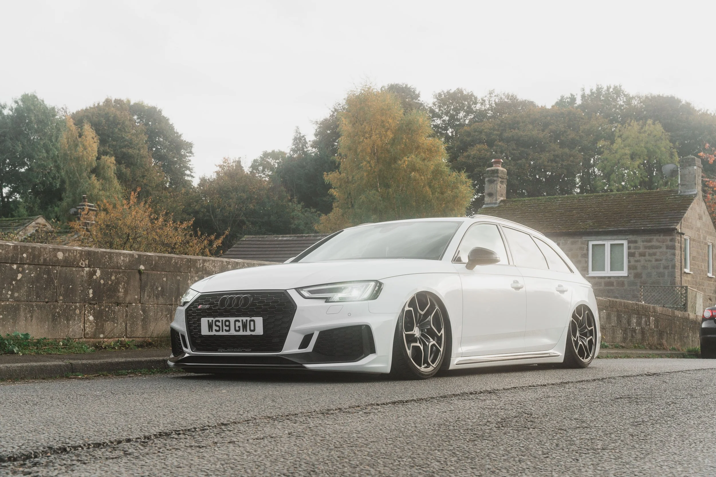 A white Audi station wagon with lowered suspension parked on the street in front of a stone wall and a house, with trees in the background.