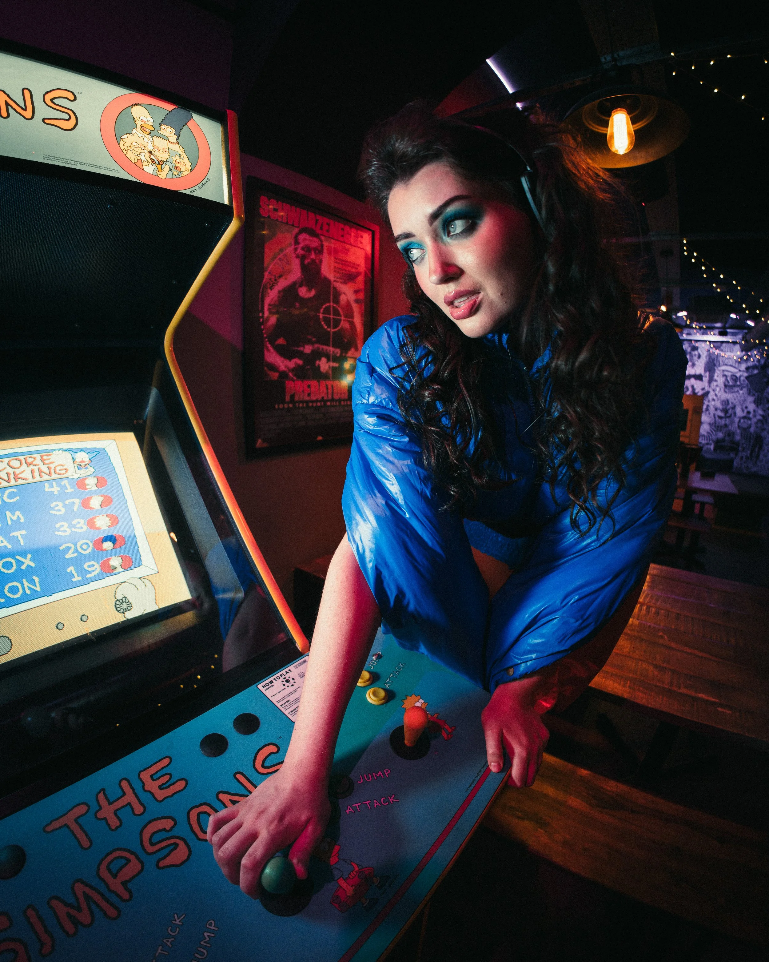 A woman with dark curly hair and blue eye makeup playing an arcade game in a dimly lit room with posters on the wall.