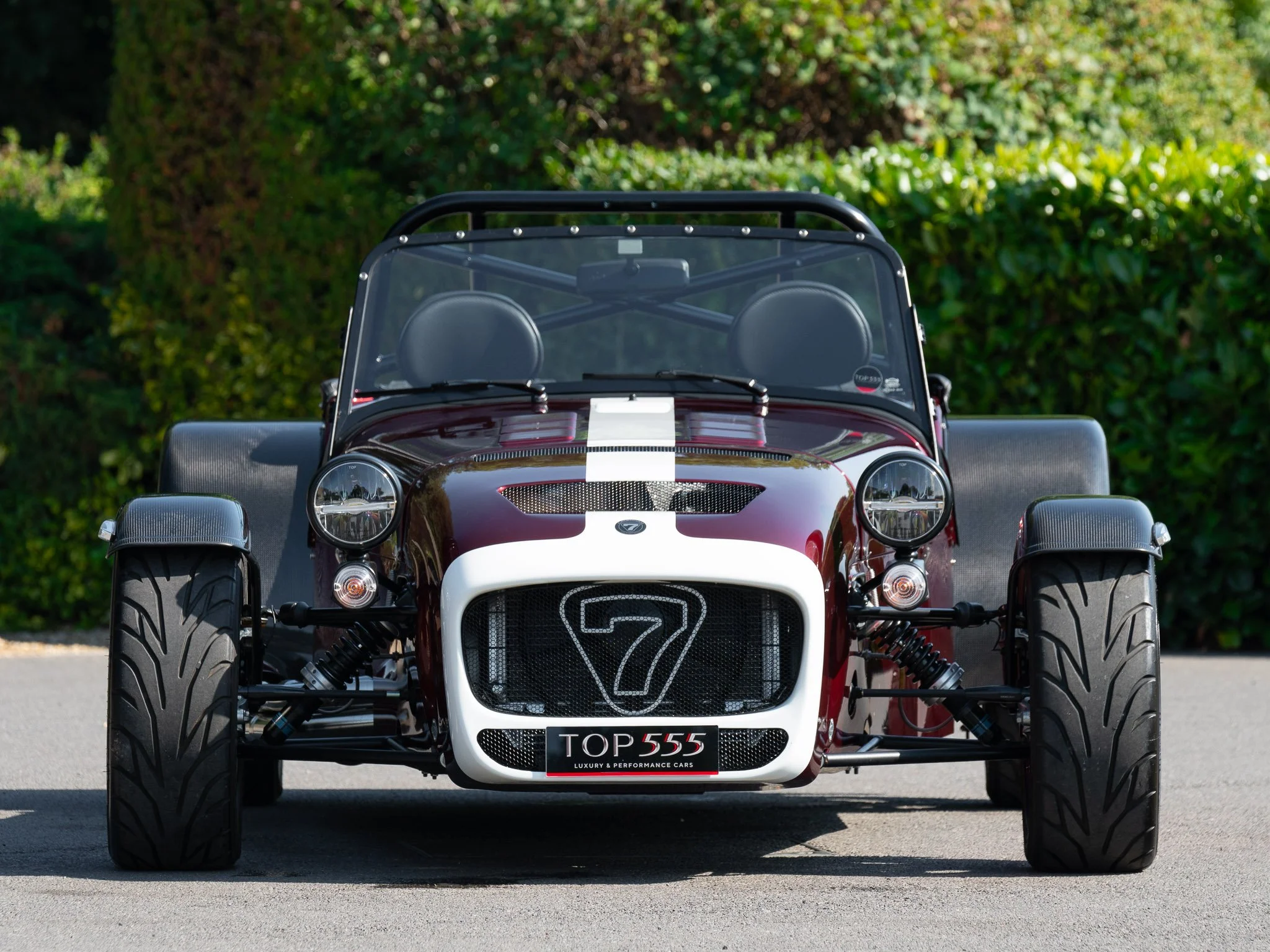 Front view of a vintage race car with a maroon body, white racing stripe, large tires, and a racing number '7' on the front grille, parked on a paved surface with greenery in the background.
