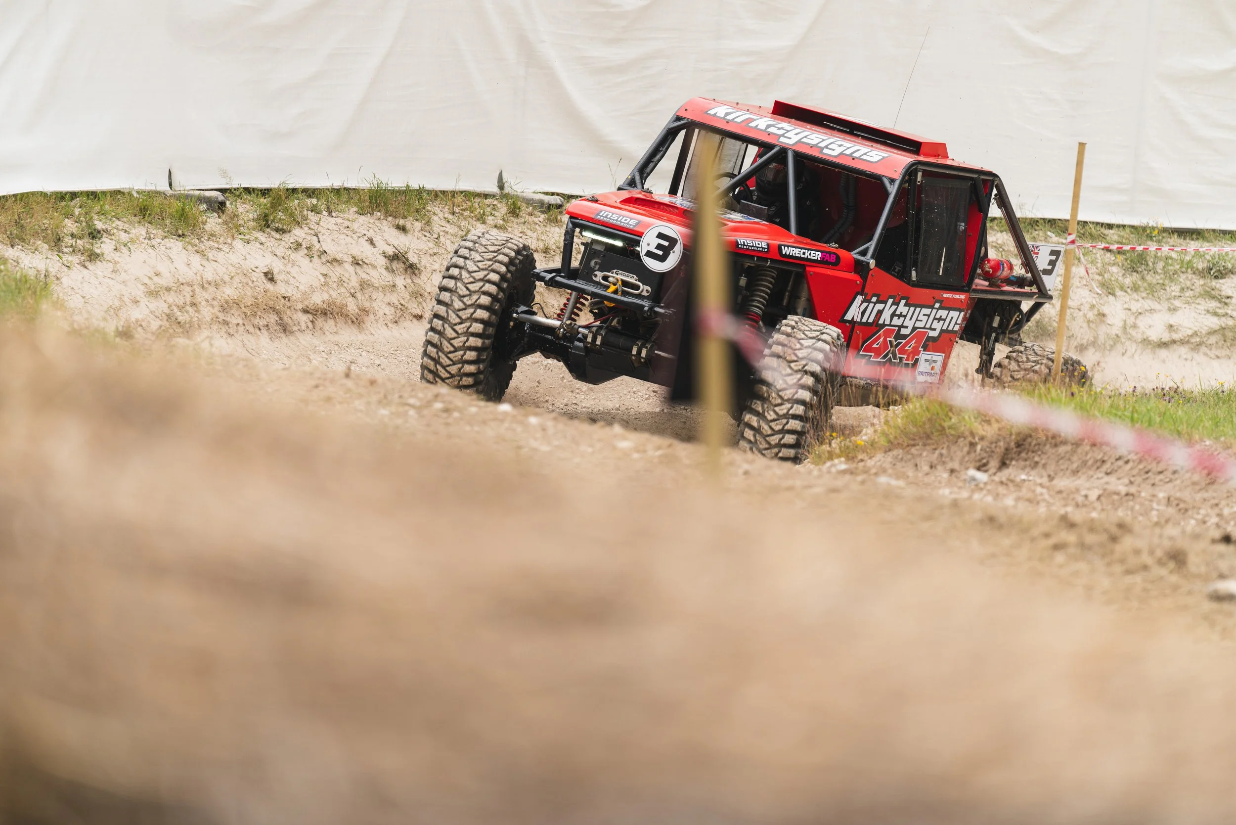 Red off-road race vehicle driving on a dirt track, surrounded by grass and a white barrier in the background.