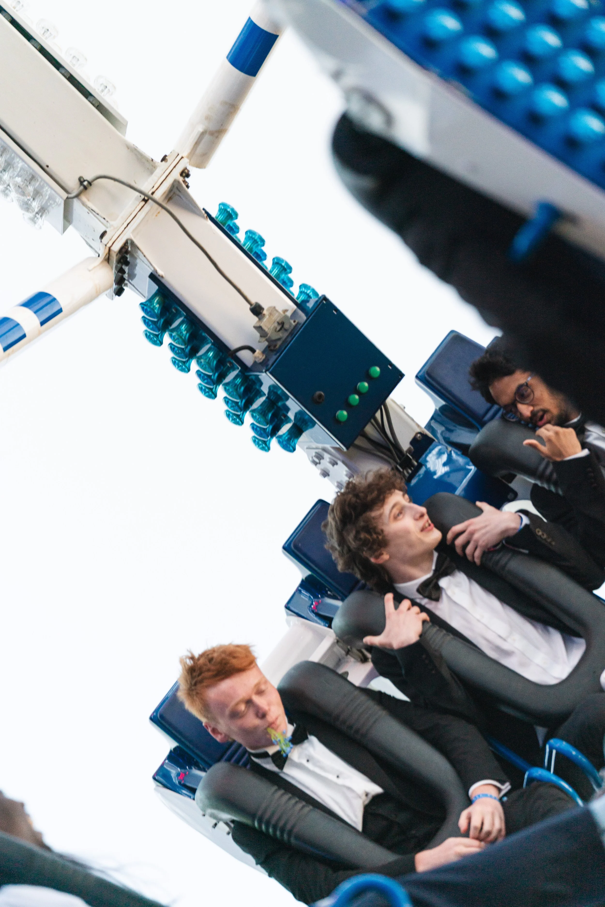 Young men in formal attire seated in amusement park ride seats, with one talking on a phone, as the ride is about to start.
