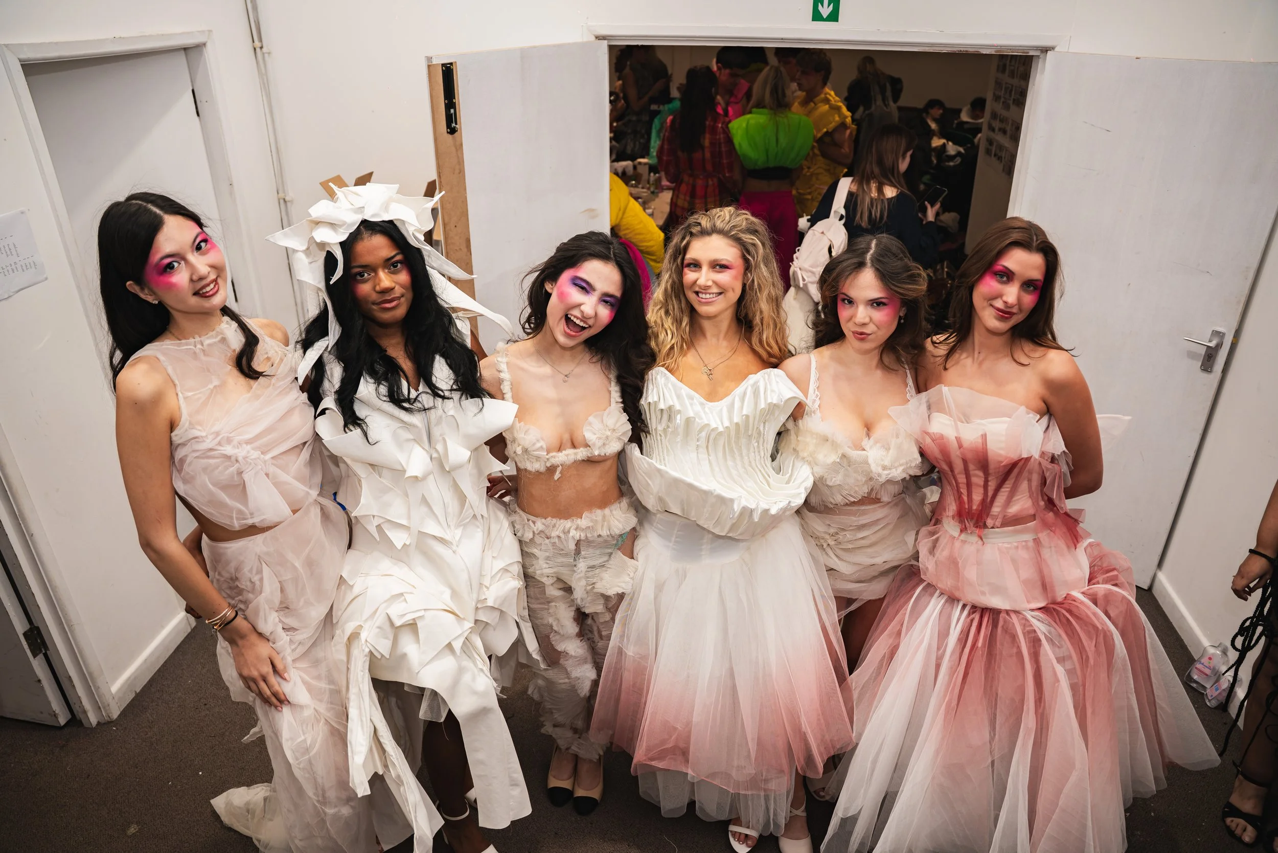 Group of six women dressed in avant-garde, playful, and colorful fashion, some with exaggerated makeup and hairstyles, posing backstage at a fashion show.