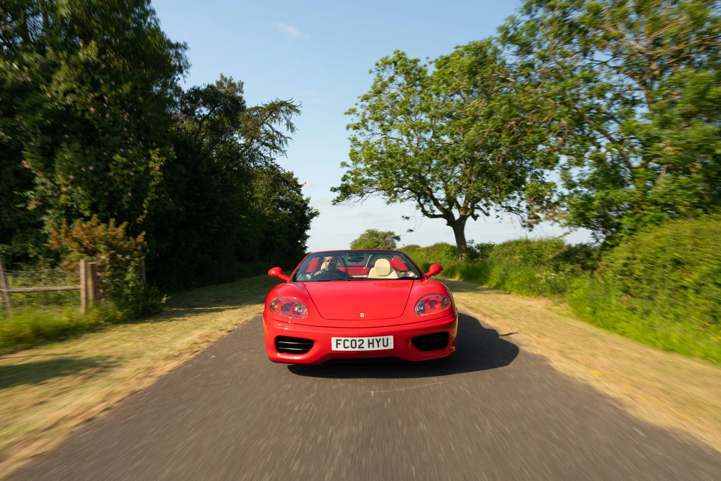 A red convertible sports car driving on a rural road with green trees and bushes on both sides under a clear blue sky.