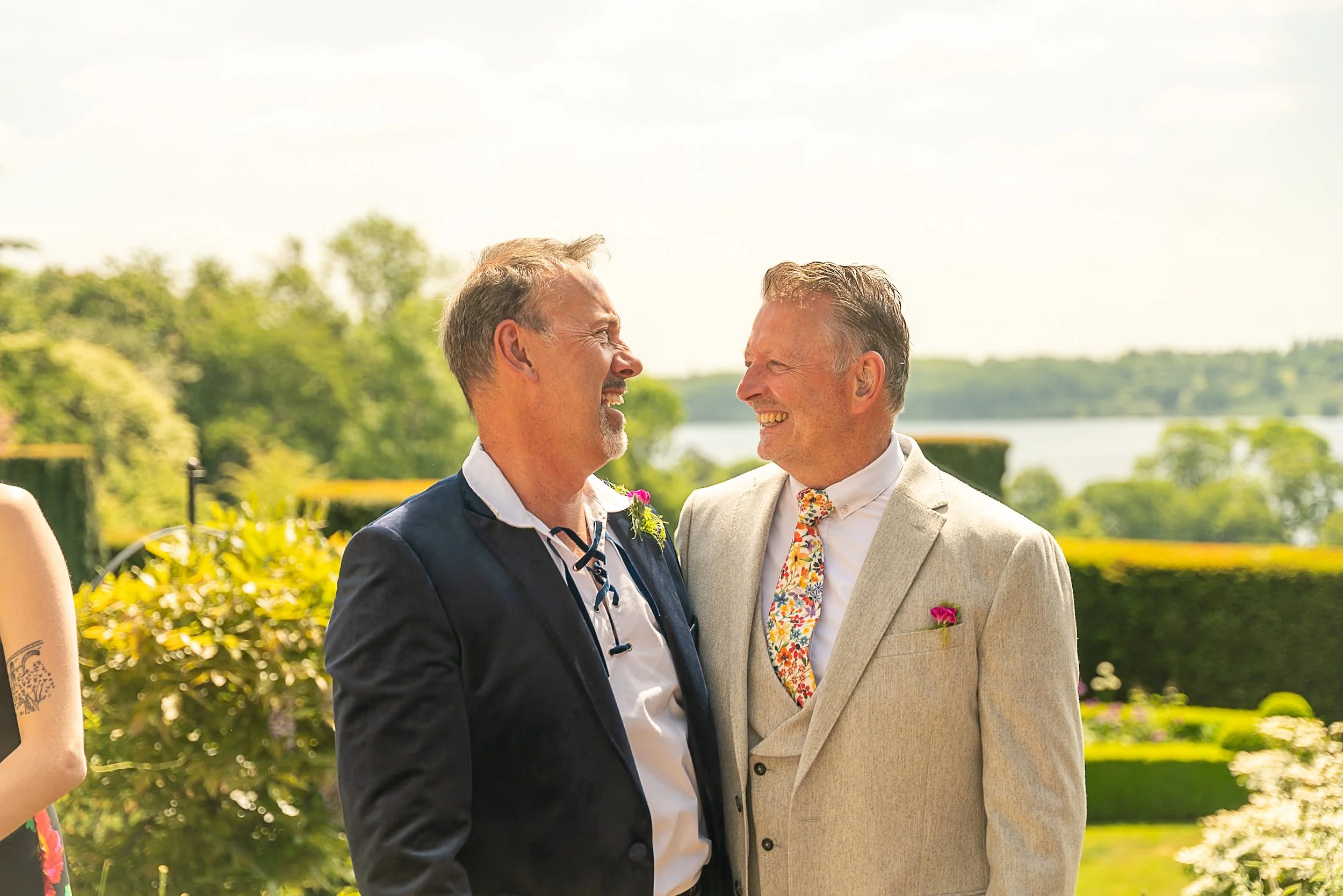 Two men in formal suits smiling and looking at each other outdoors during a wedding celebration with green trees and water in the background.