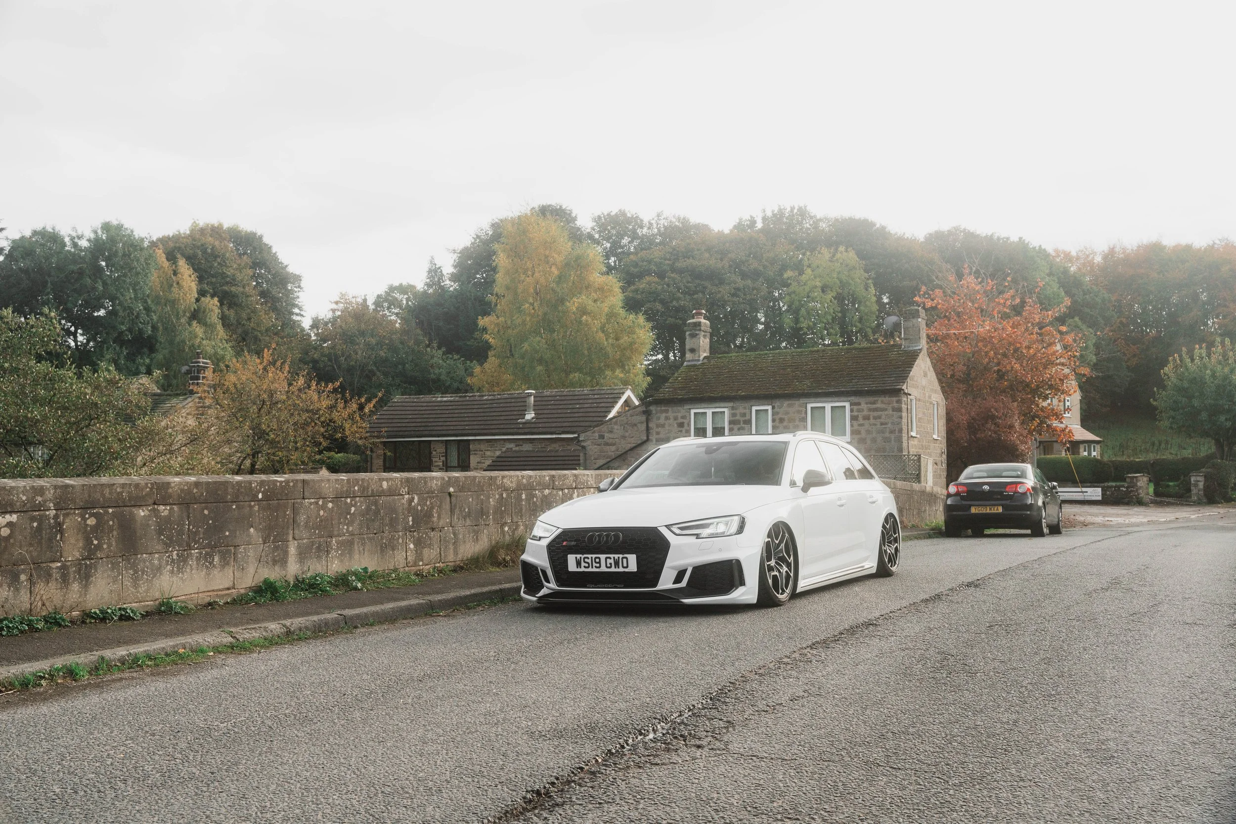 A white Audi car parked on a residential street with a stone wall and several other cars, surrounded by trees with autumn foliage.