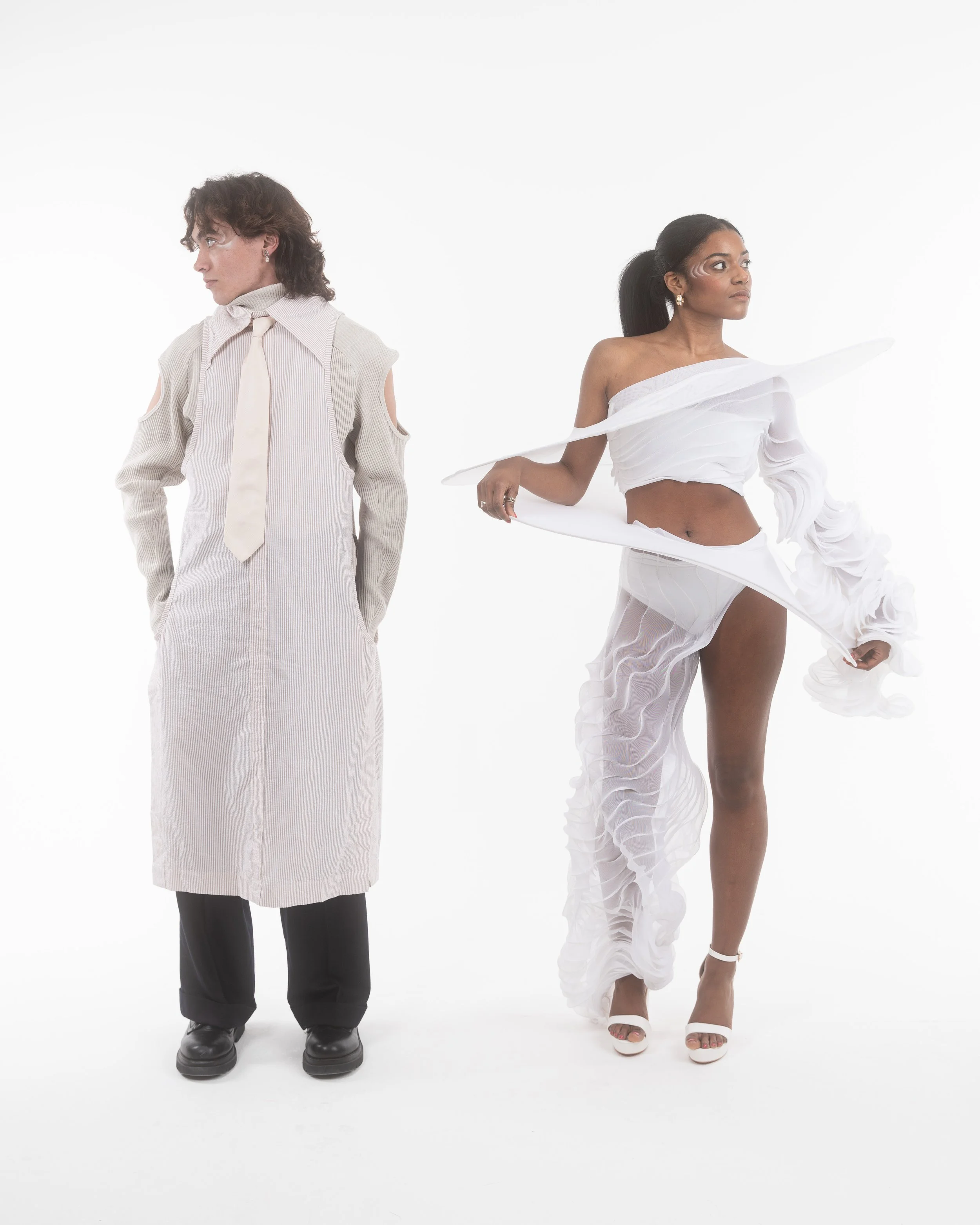 Two people standing against a white background, one man in a beige trench coat and the other woman in a white designer outfit.