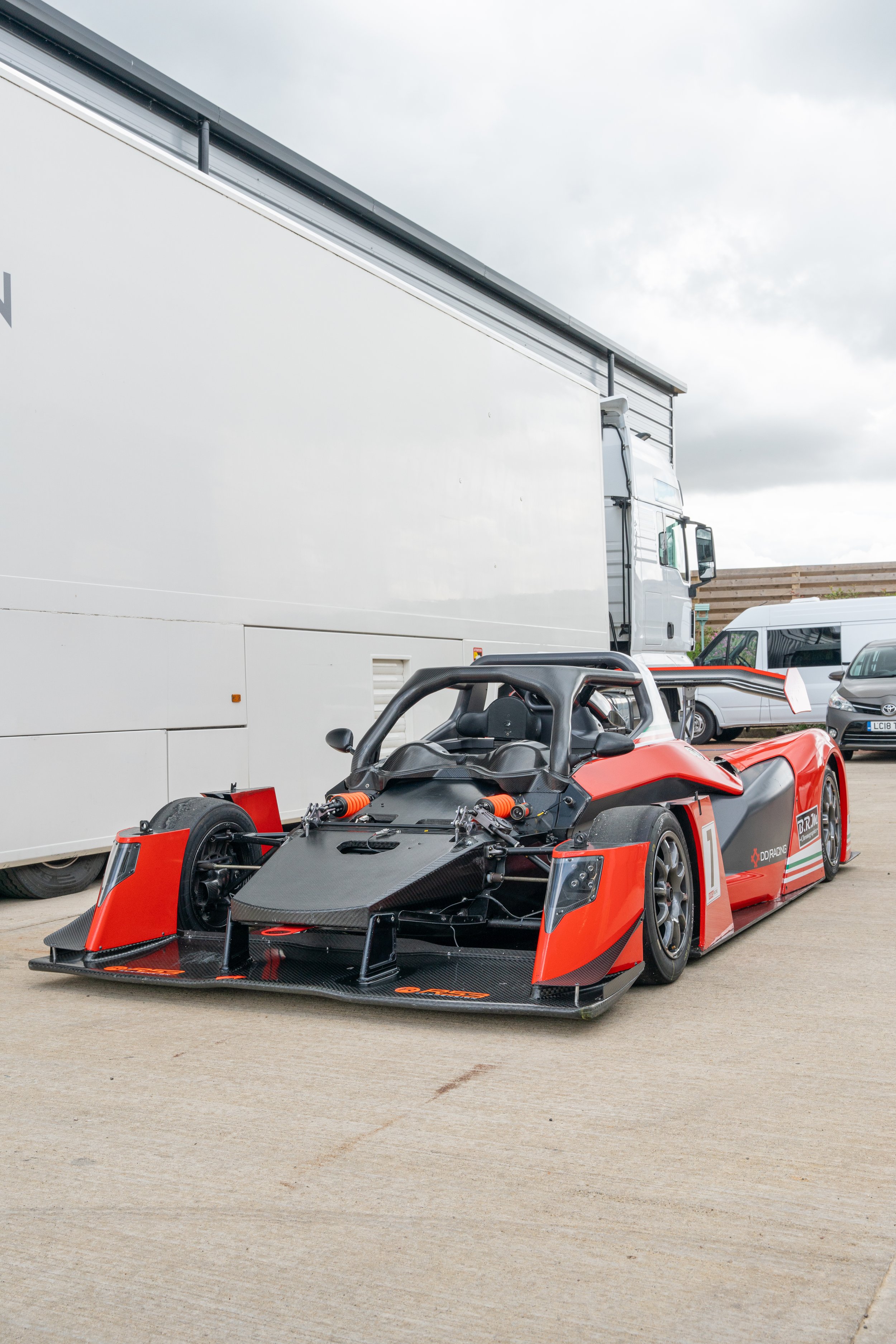 A racing car with a black and red body parked beside a large white truck, under a cloudy sky.