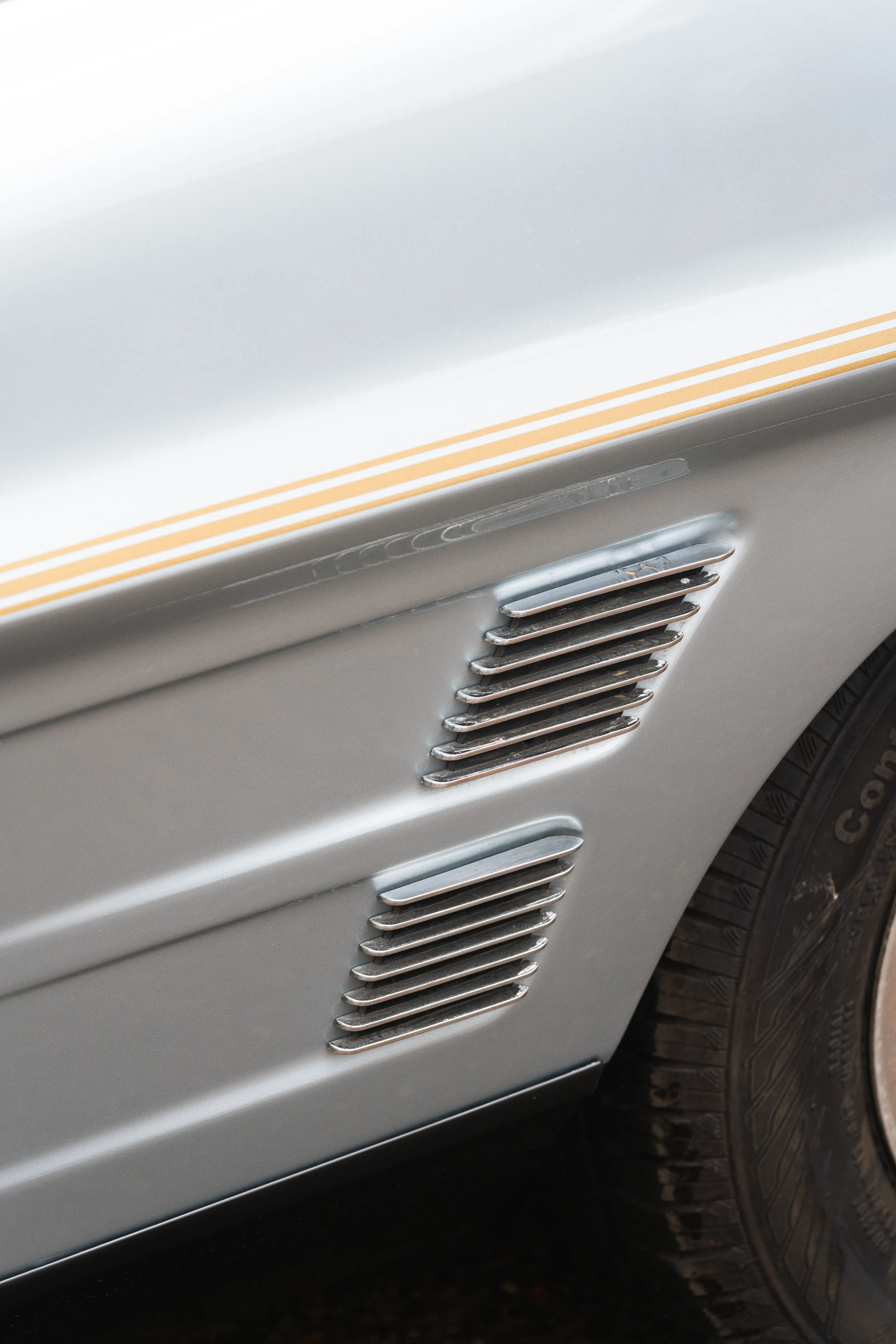 Close-up of a vintage silver car's side panel with multiple horizontal chrome vents and a part of a black tire in the bottom right corner.