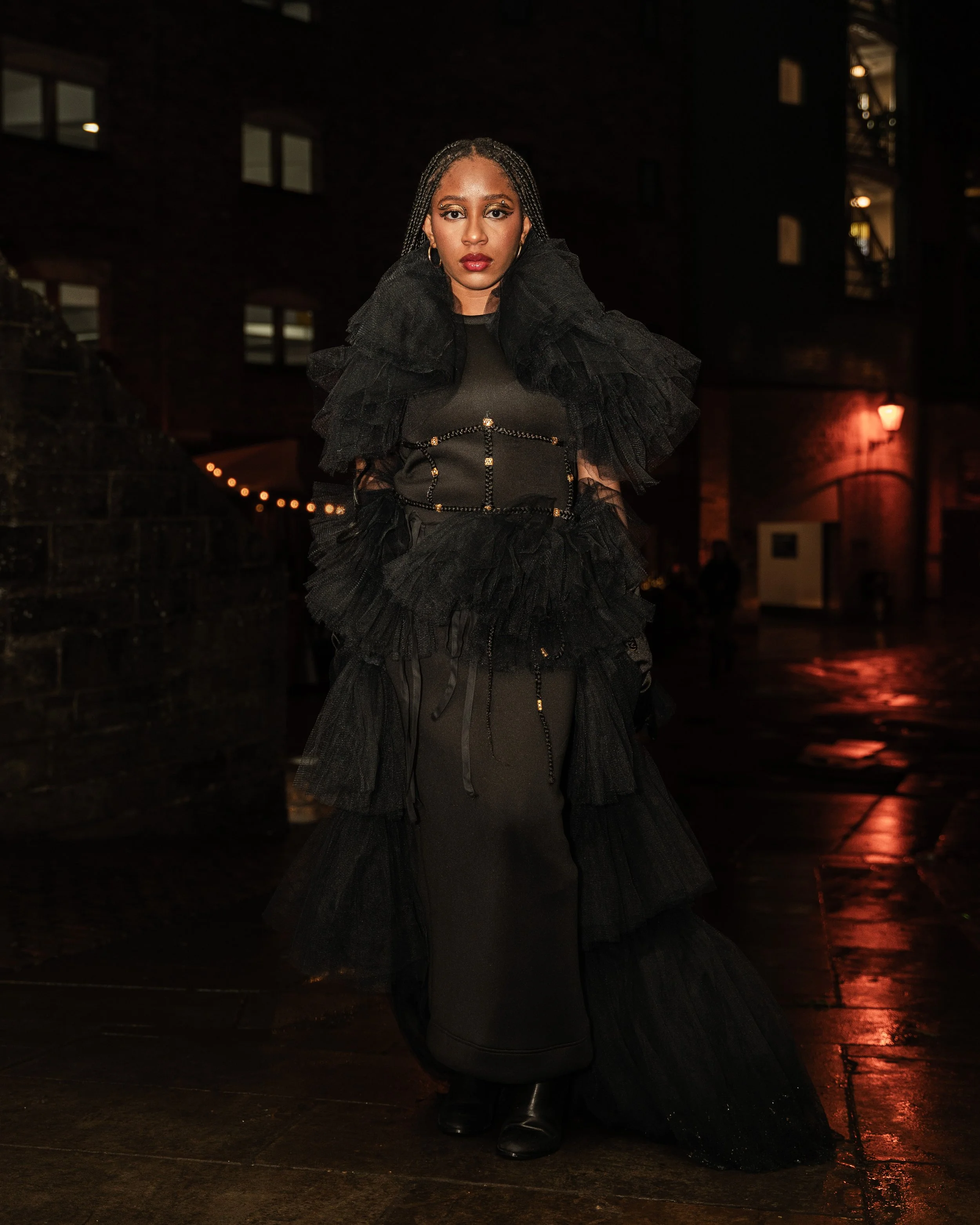 A woman standing outdoors at night dressed in a black, layered tulle and leather fashion ensemble with a dark background and dim street lighting.