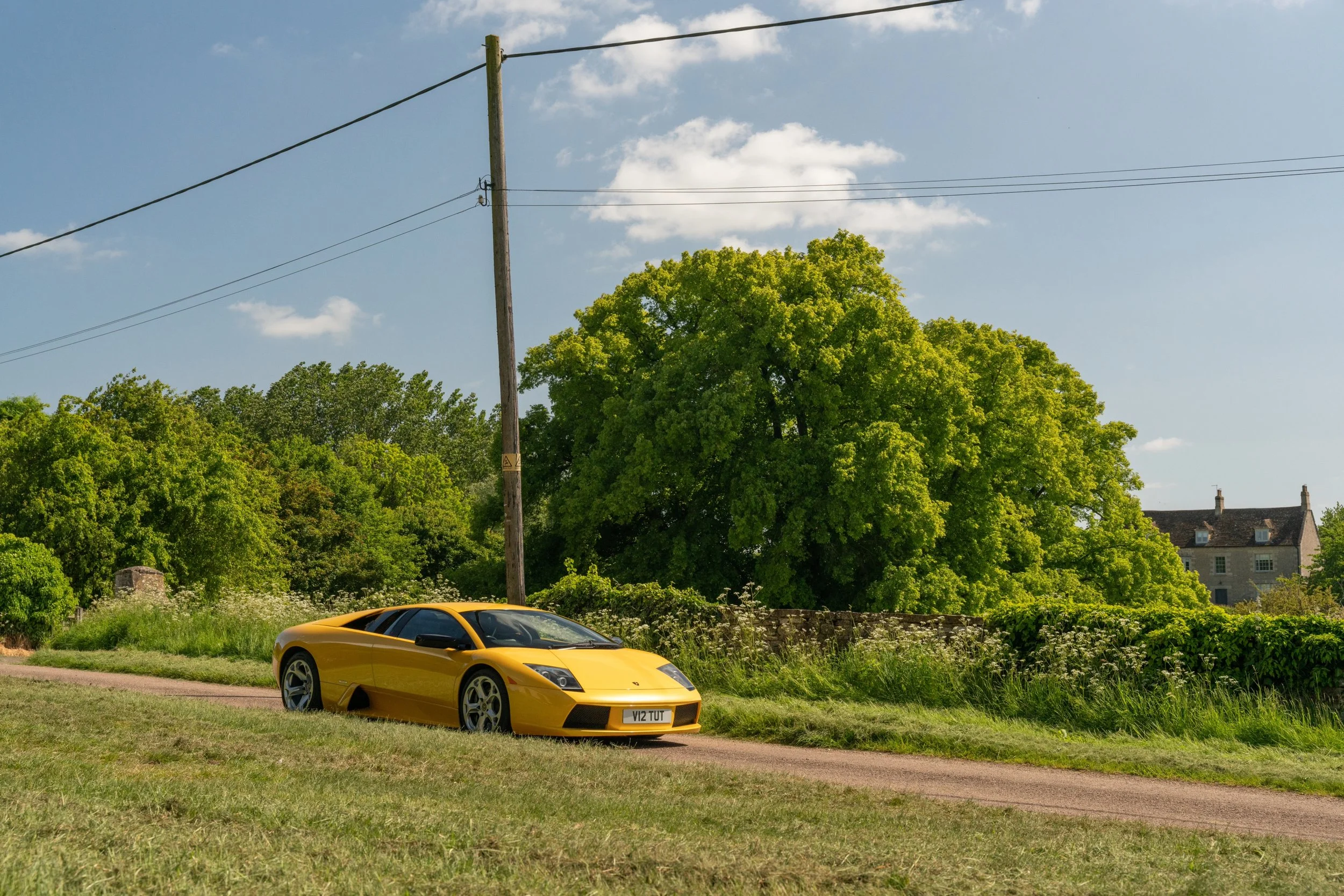 A yellow Lamborghini sports car parked on a dirt road with green trees and a house in the background.