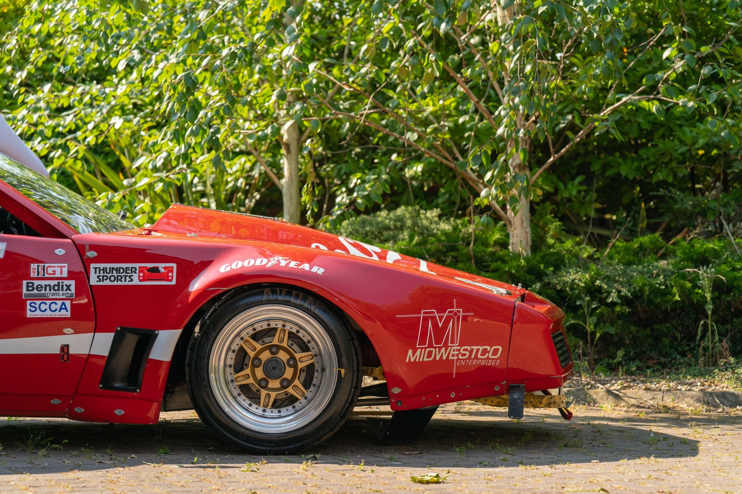 Close-up of a red race car's front left side with sponsorship logos, parked on a paved surface with green foliage in the background.
