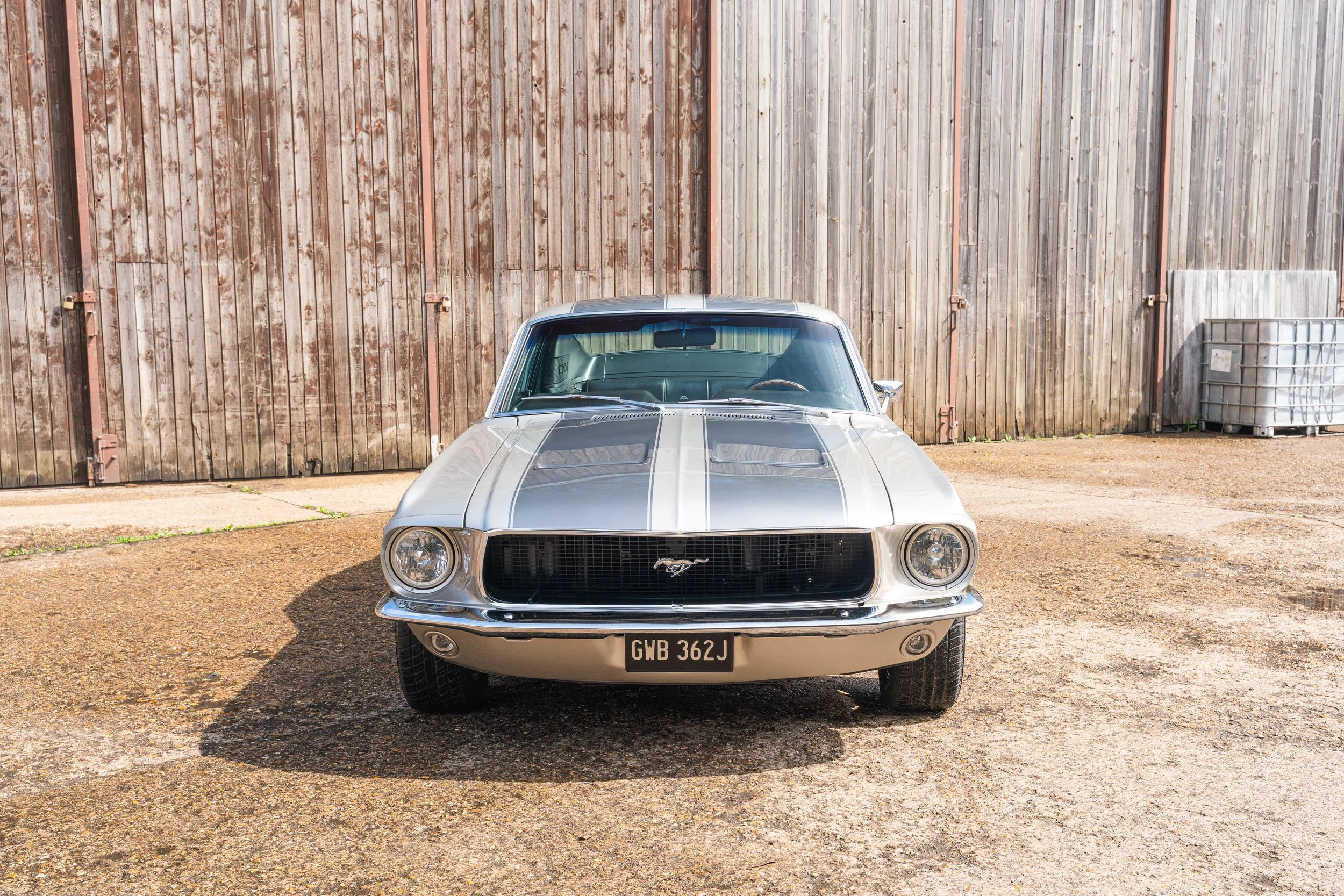 Front view of a vintage silver Ford Mustang with black racing stripes parked on a gravel surface in front of a wooden fence.