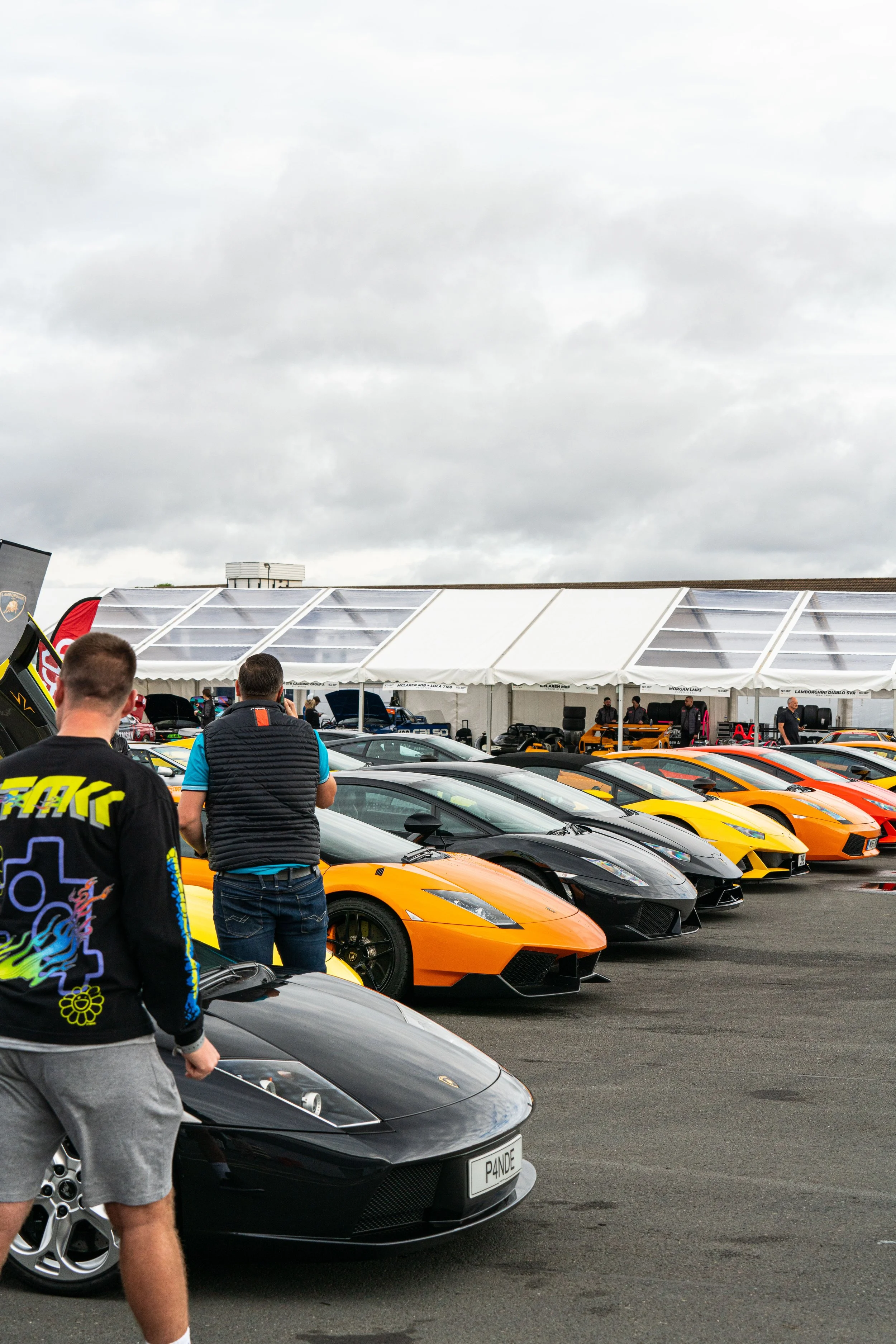 A row of colorful Lamborghini sports cars on display at an outdoor car event, with people walking and standing nearby under a cloudy sky.