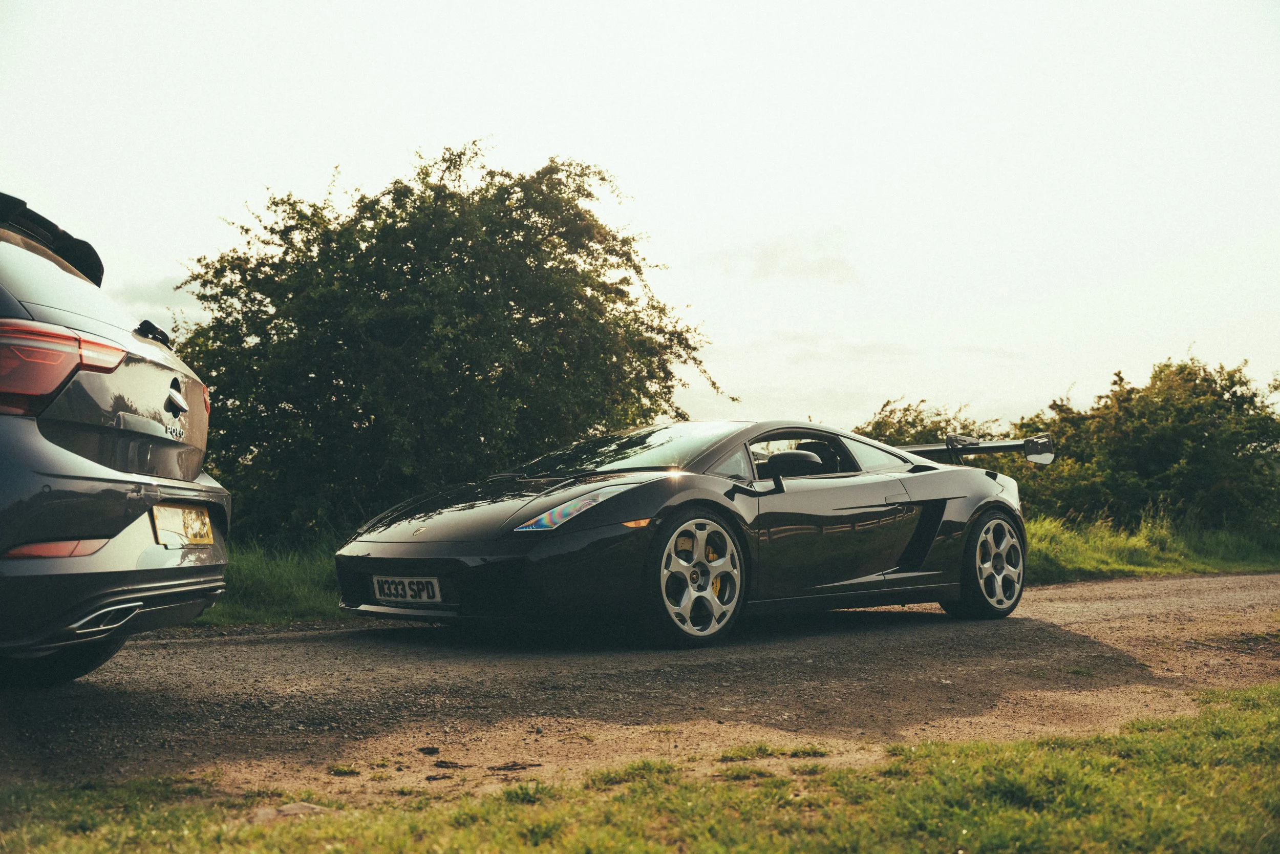 A black sports car with a rear spoiler parked on the side of a rural road next to an SUV, with greenery and trees in the background during daylight.