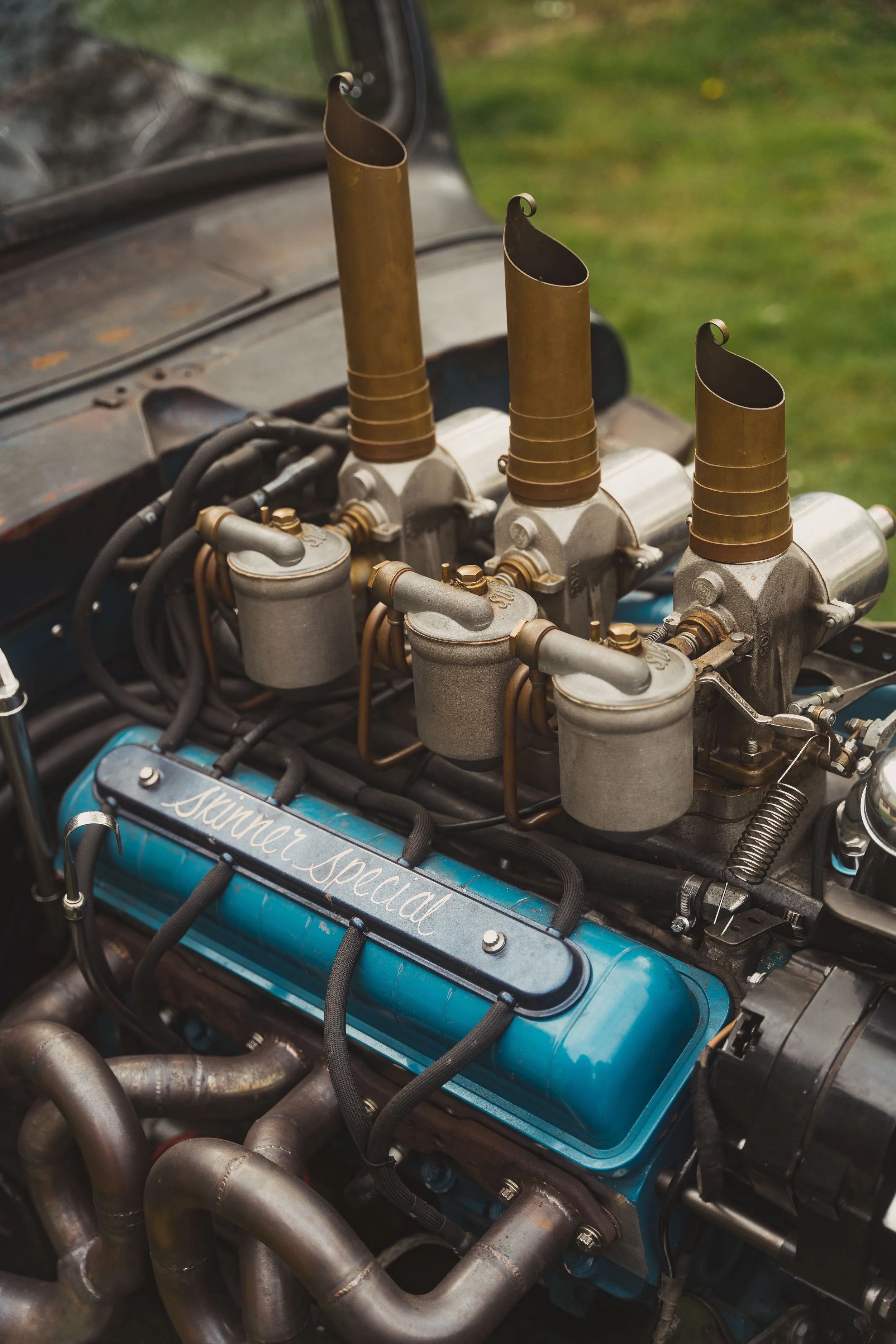 Close-up of a vintage car engine with a blue valve cover labeled 'Shaner Special,' four carburetors with brass-colored intake stacks, and various metal pipes and hoses.