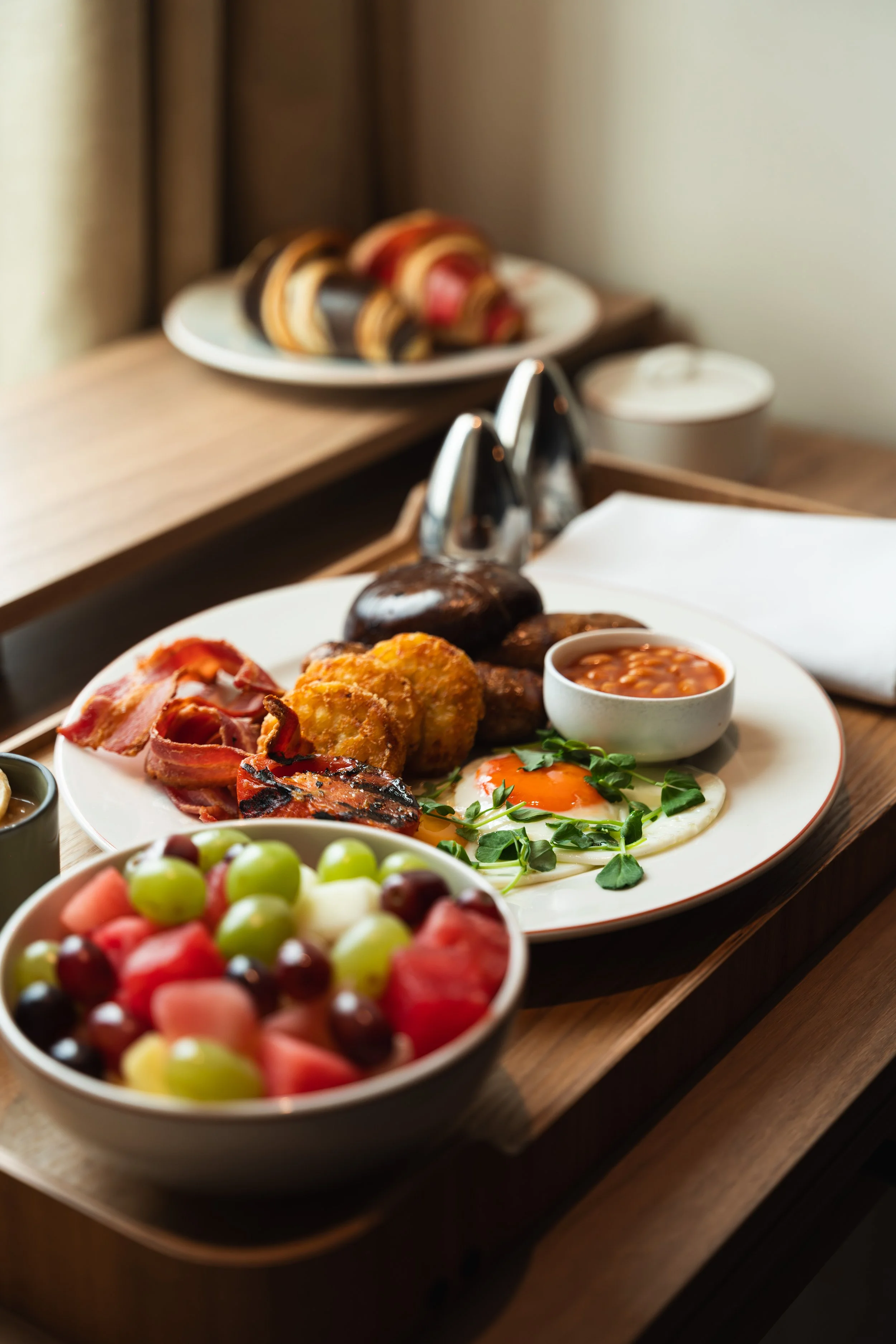 A breakfast plate with eggs, bacon, hash browns, sausage, and baked beans, with a bowl of mixed fruit in the foreground and a fruit pastry on a plate in the background.