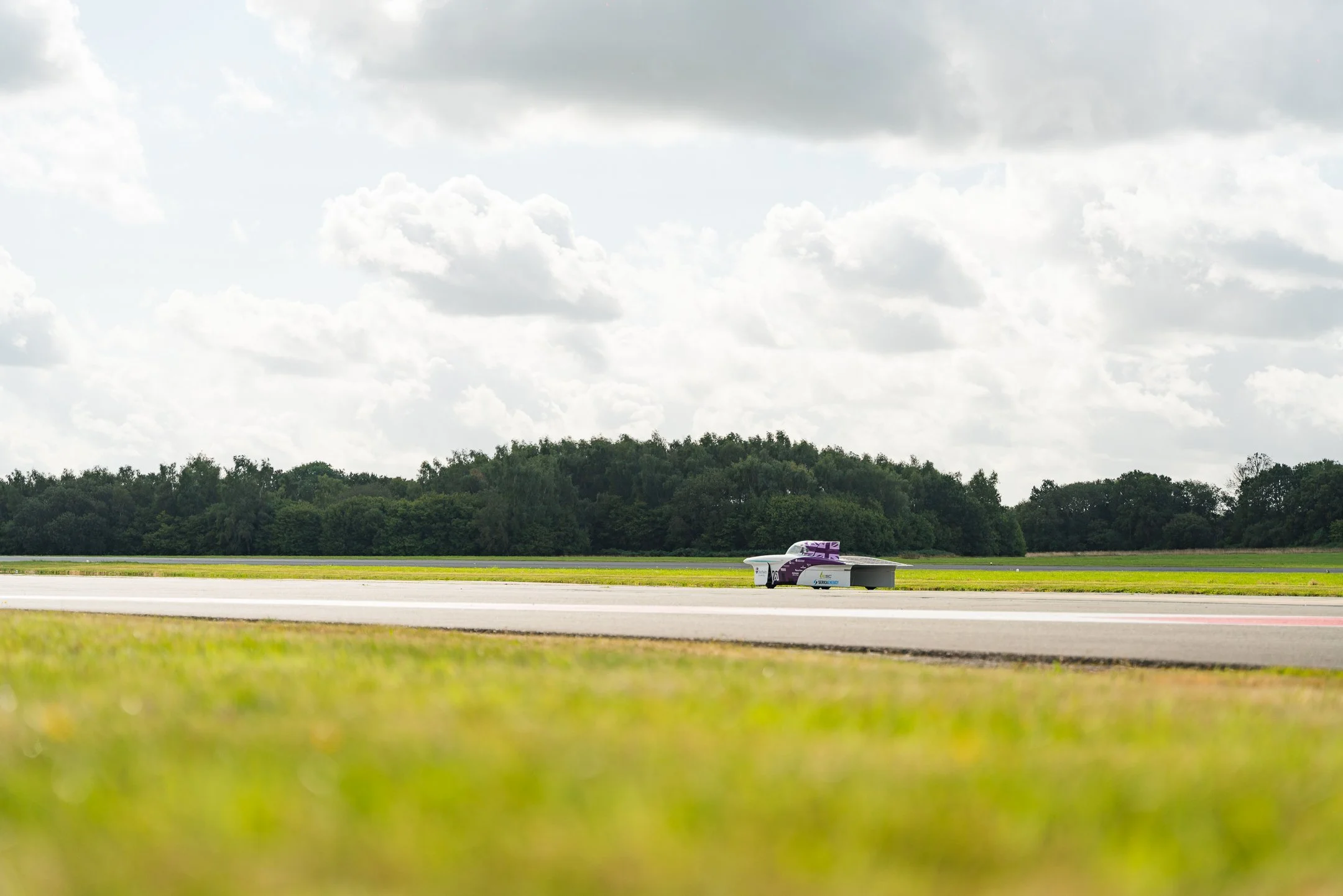 A sleek, futuristic racing vehicle on a runway with a backdrop of trees and a cloudy sky.