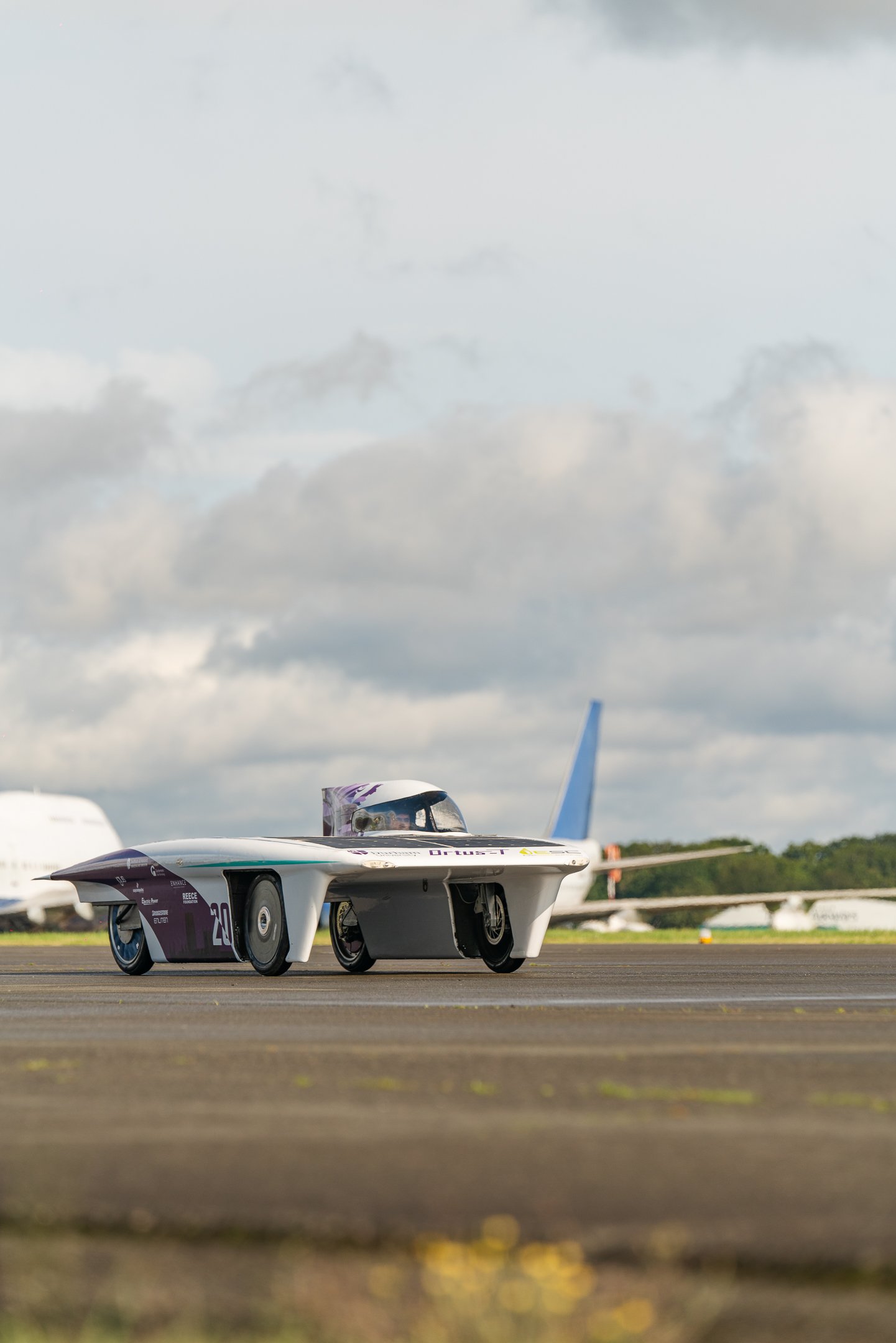 A futuristic, sleek racing vehicle on an airstrip, with a modern airplane in the background under cloudy skies.