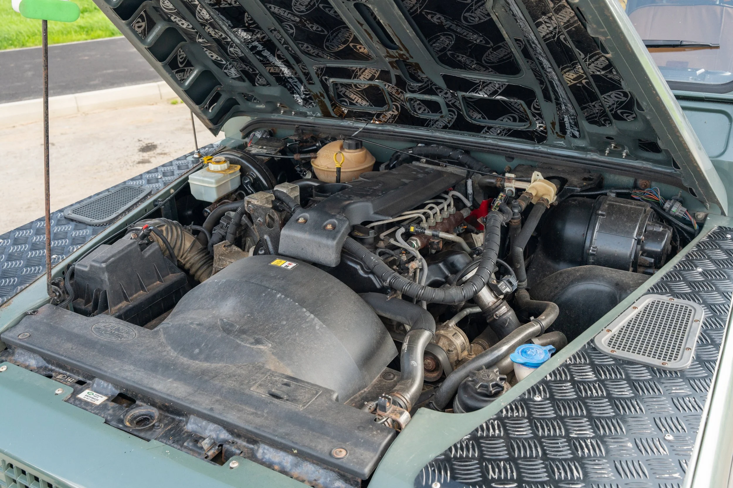 Open vehicle hood revealing engine bay with various components, hoses, and wiring, with a diamond plate pattern on the surrounding metal surfaces.
