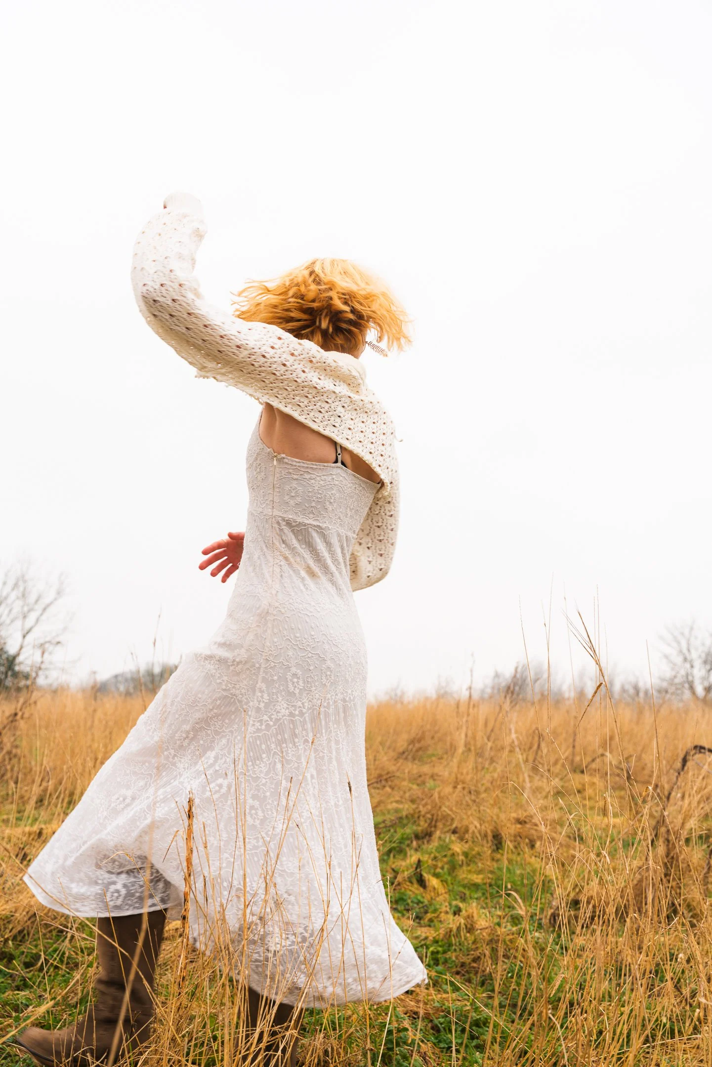 A woman with curly hair wearing a white lace dress and a knitted cardigan, standing in a field of dry grass, with her arm raised as her hair blows in the wind, against a cloudy sky.