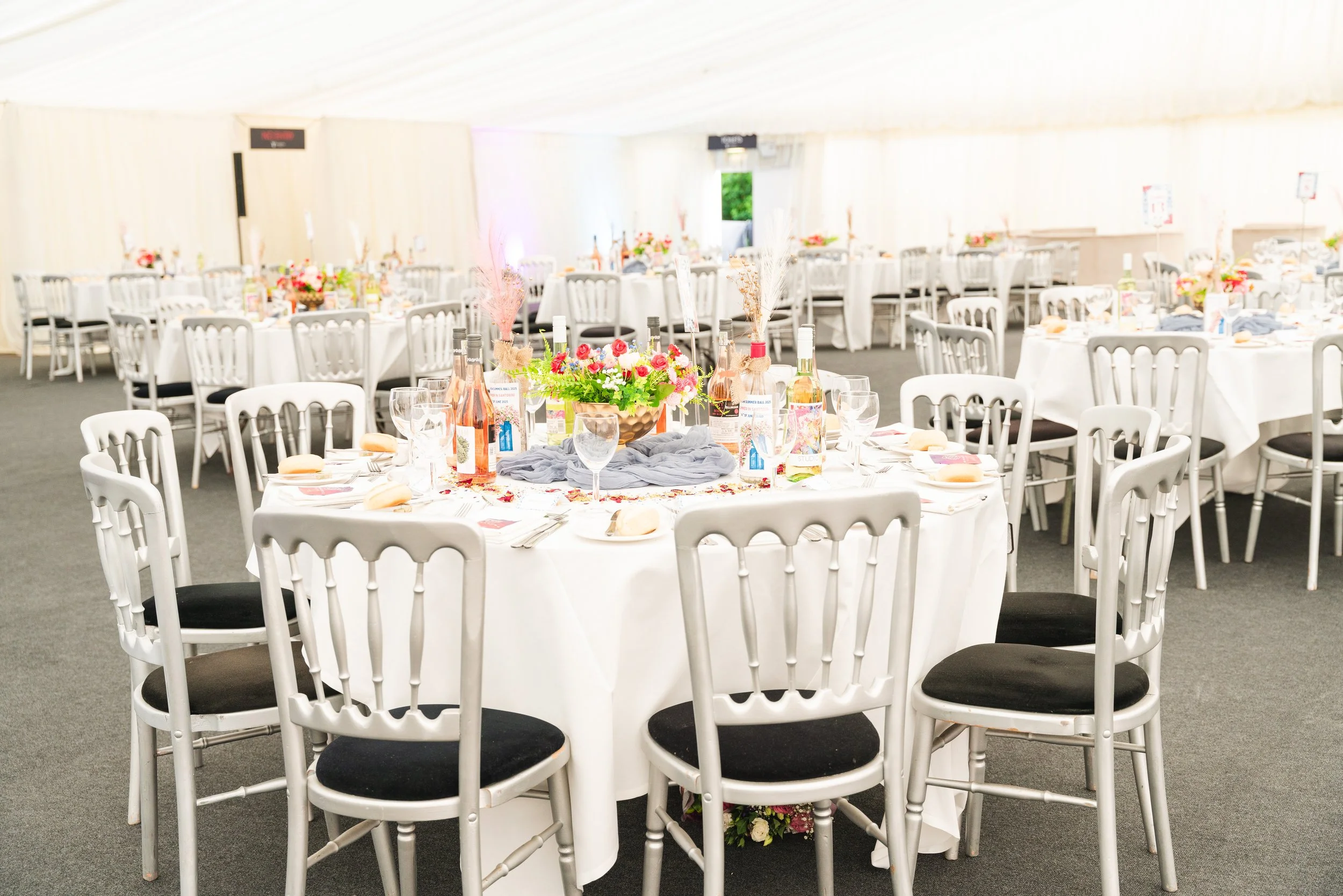 Wedding reception venue with round tables covered in white tablecloths, decorated with floral centerpieces and set with plates, glasses, and utensils, inside a large white tent.