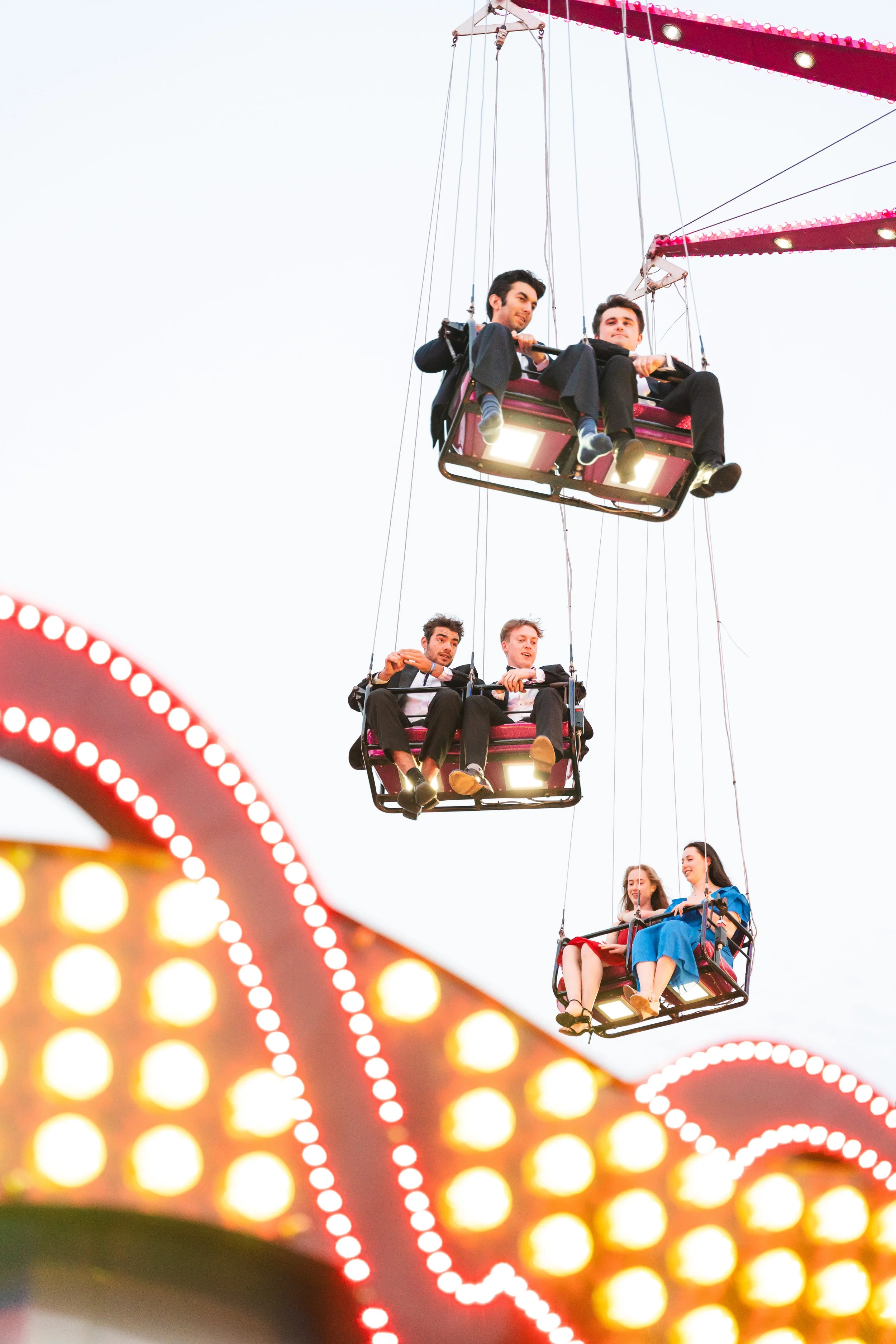 Three pairs of people riding amusement park swings, with a brightly lit ride in the foreground and a twilight sky in the background.