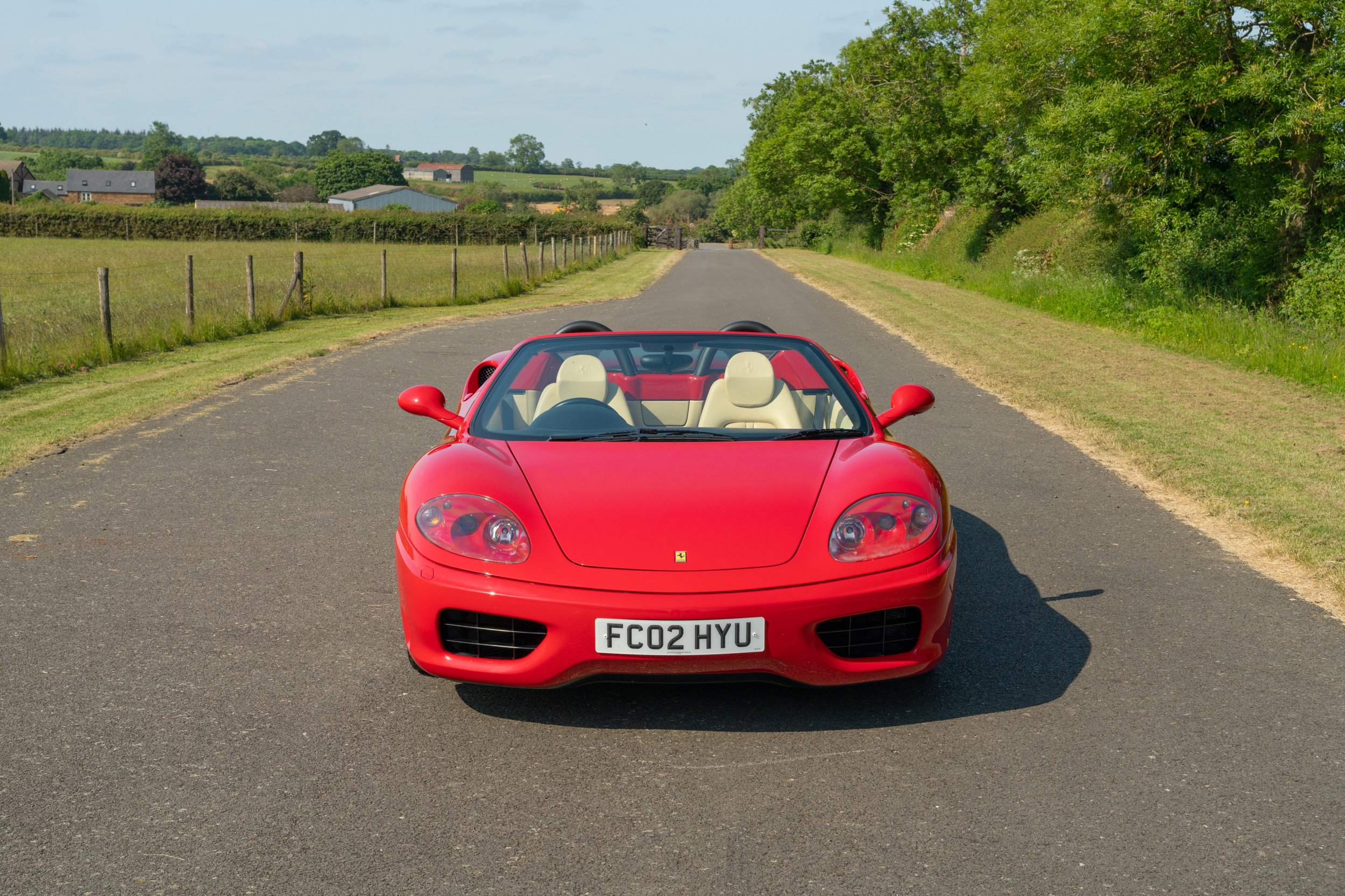 A red Ferrari convertible sports car parked on a rural road with green fields and trees in the background.