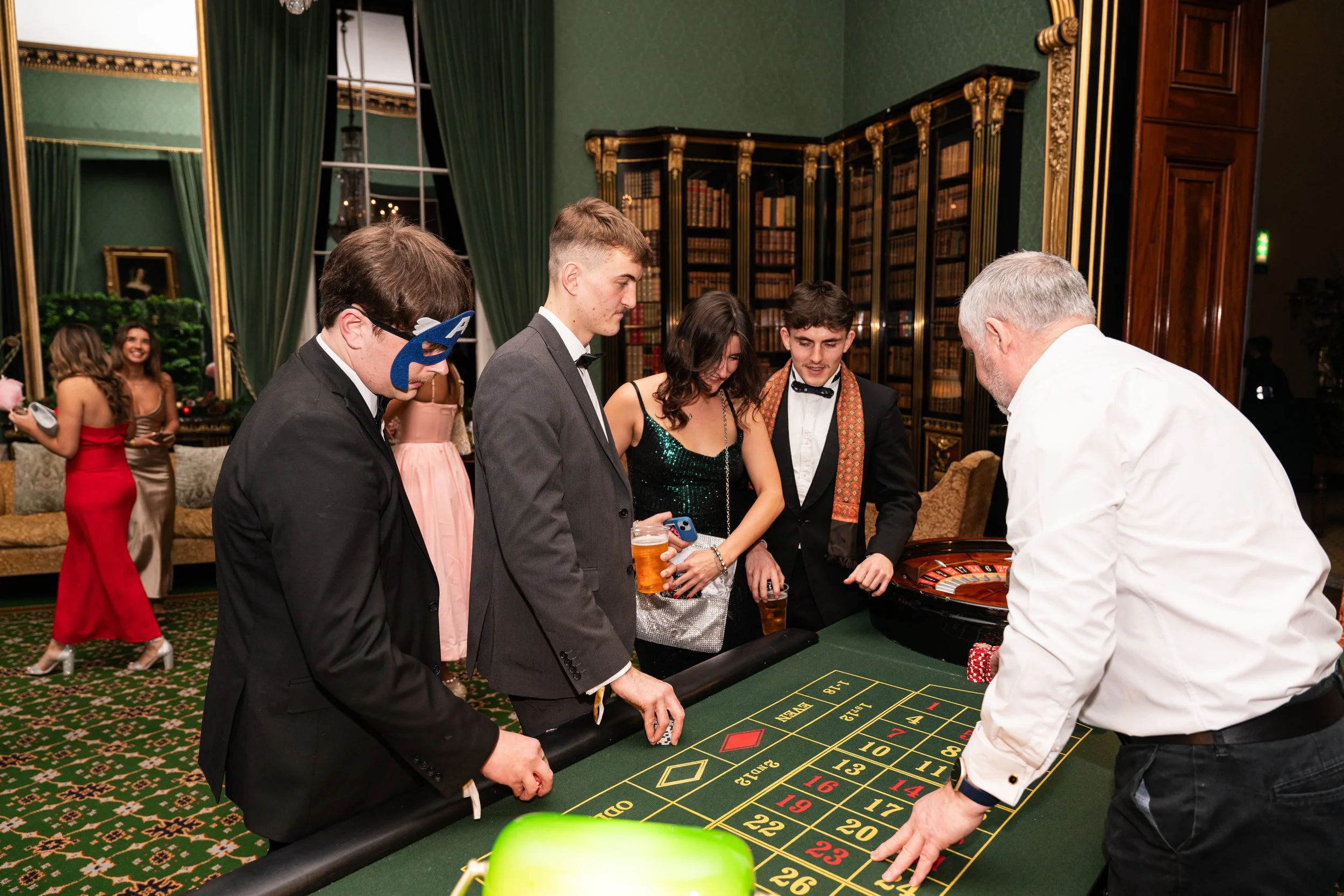 People playing roulette at a casino celebration, dressed in formal attire, with some wearing masks, and holding drinks.