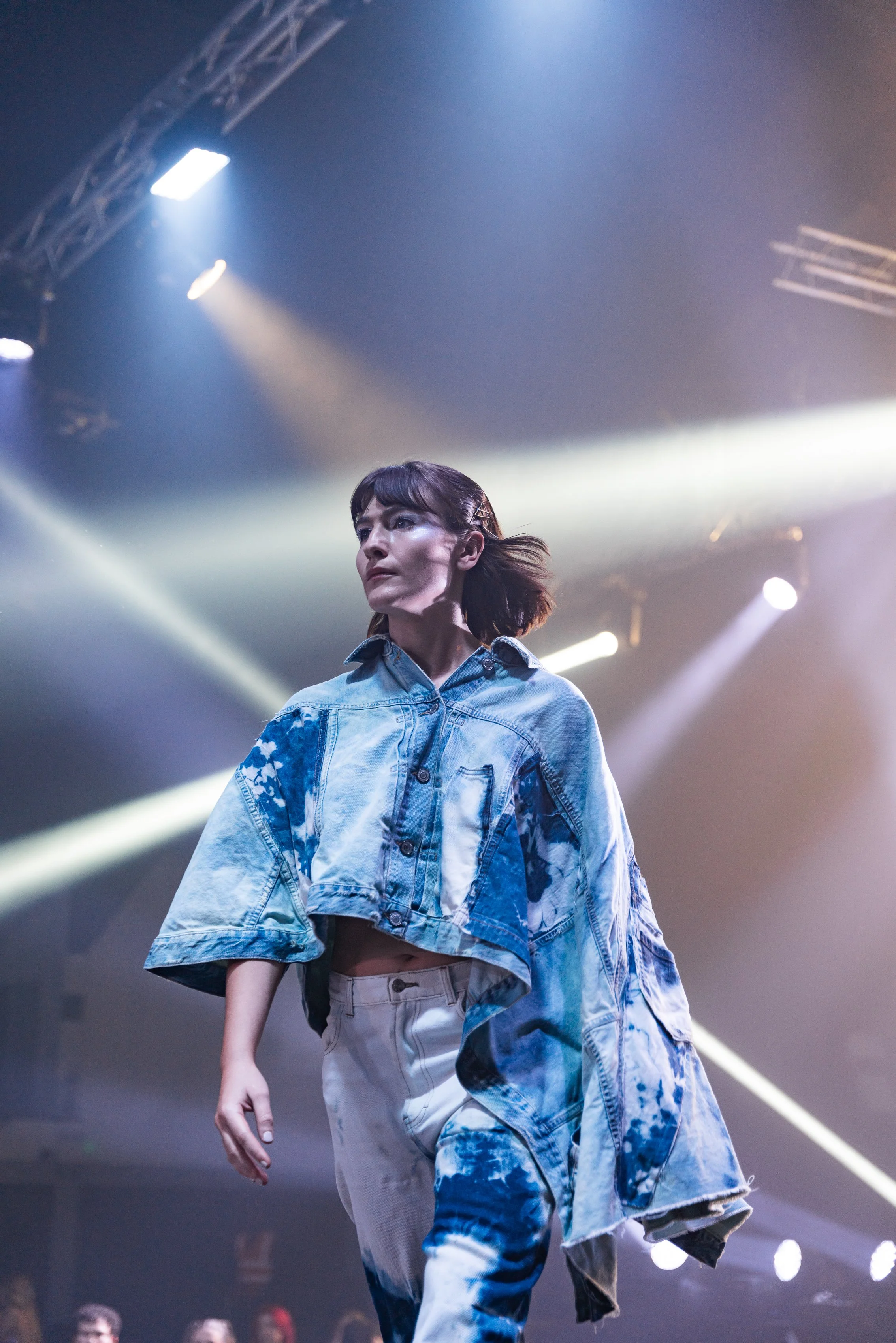 Female model walking runway in denim outfit with blue and white tie-dye pattern under stage lights.
