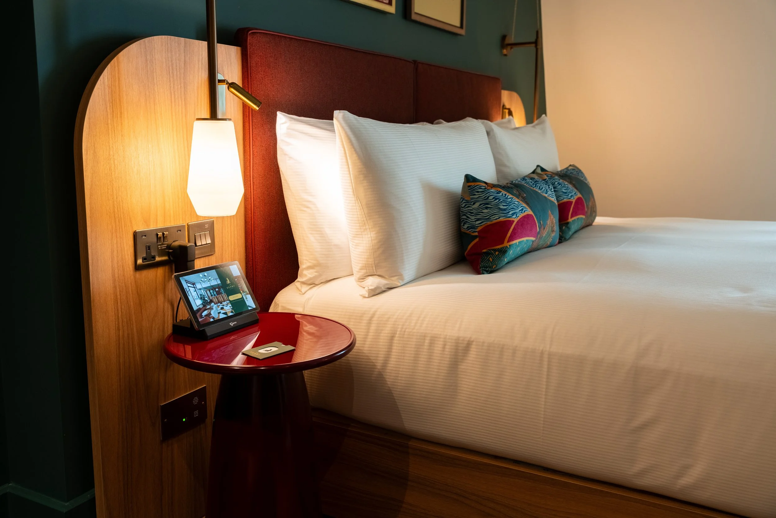 A neatly made hotel bed with white pillows and decorative pillows featuring colorful patterns, a wooden headboard, a bedside lamp attached to the headboard, a red round side table with a digital device on it, electrical outlets and switches on the he