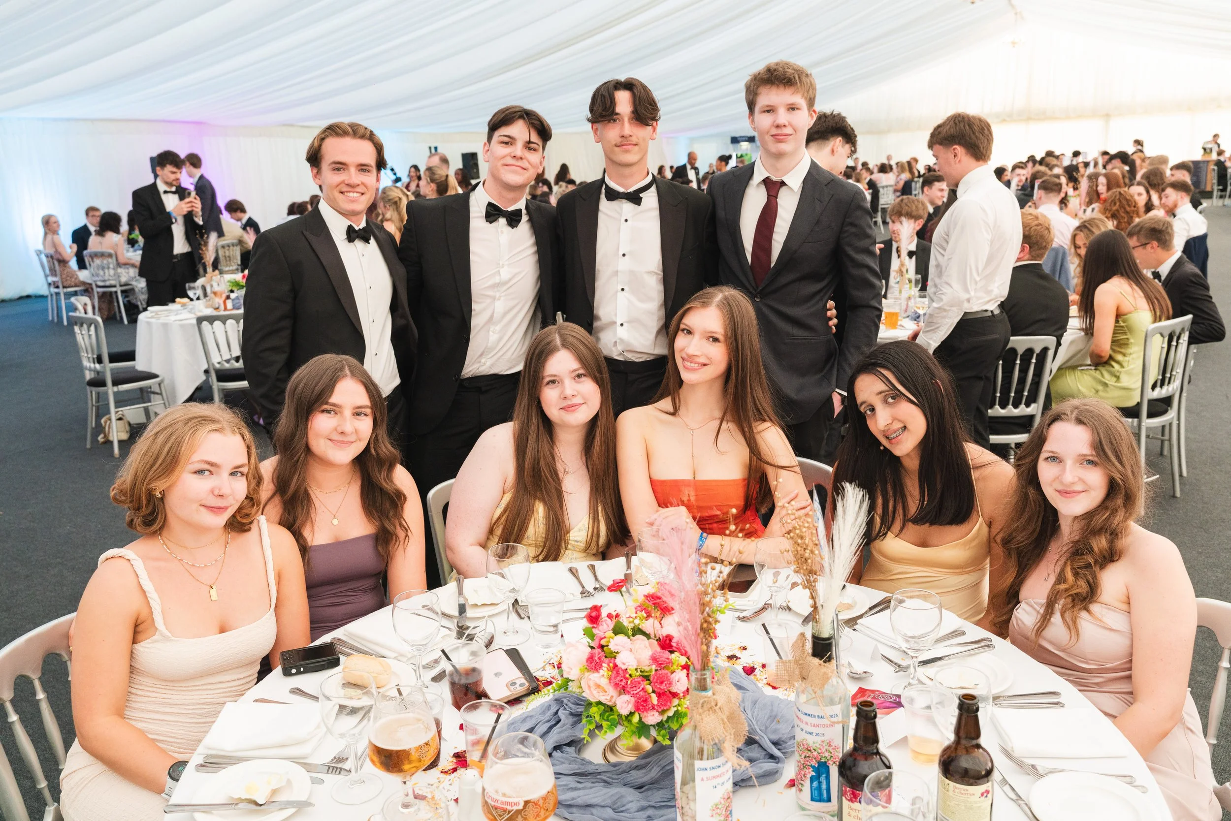 Group of young people celebrating at a formal event, with men in tuxedos standing behind women in elegant dresses sitting at a decorated banquet table.