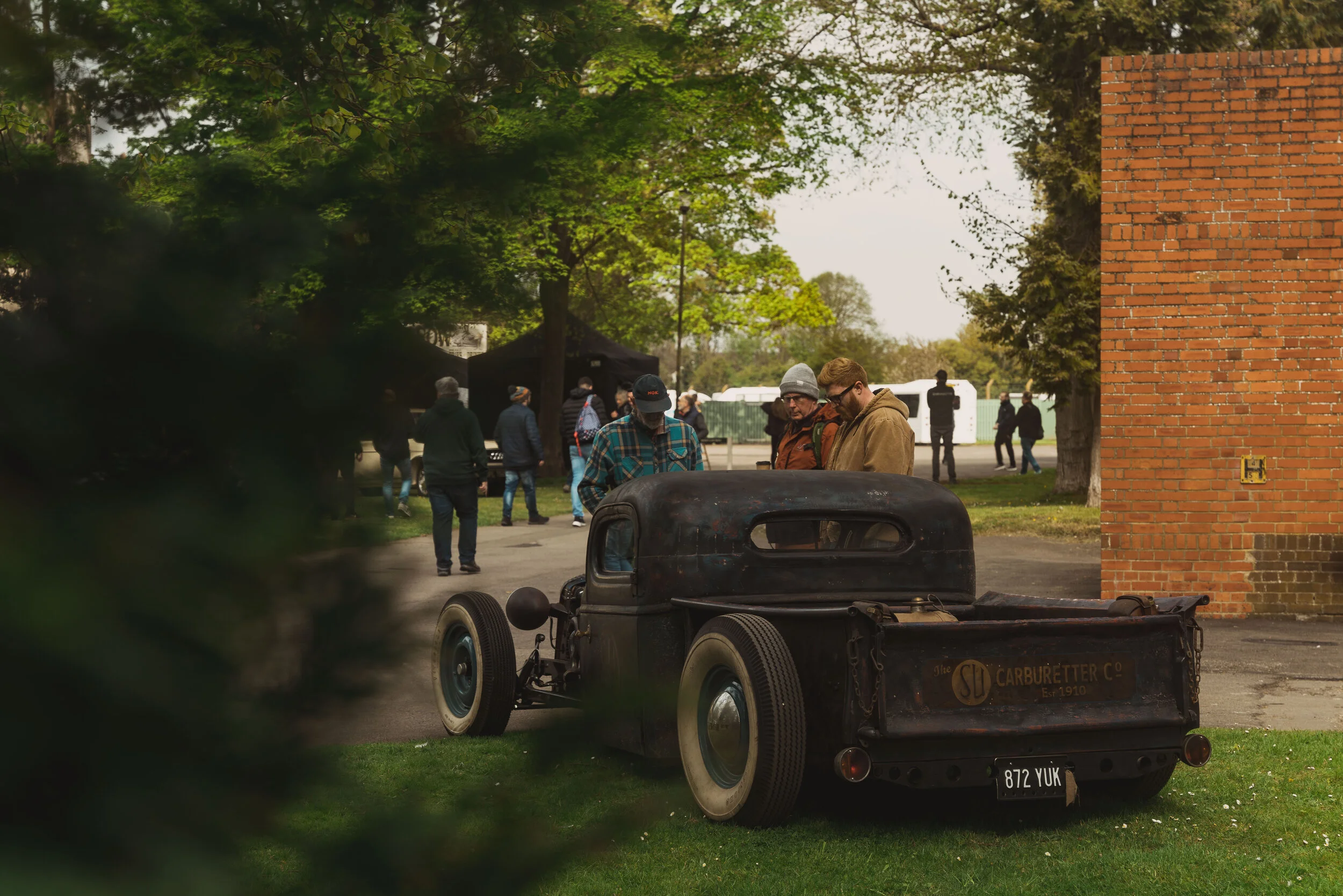 Vintage black truck with a sign for The Carburetter Co. on the back, parked on green grass, with three men examining it, outside on a cloudy day with trees and people in the background.