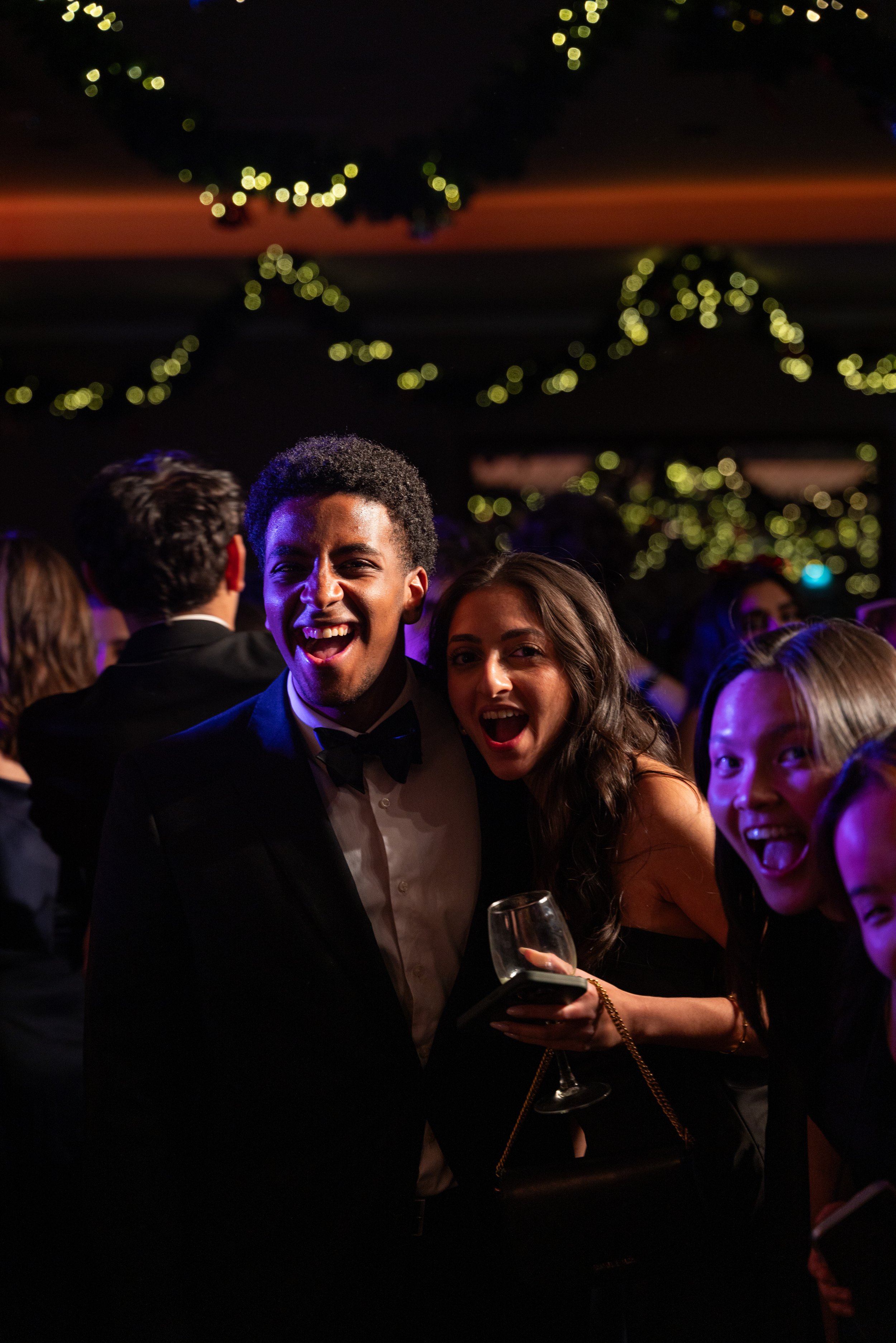 People dressed in formal attire celebrating at a party with festive string lights in the background.