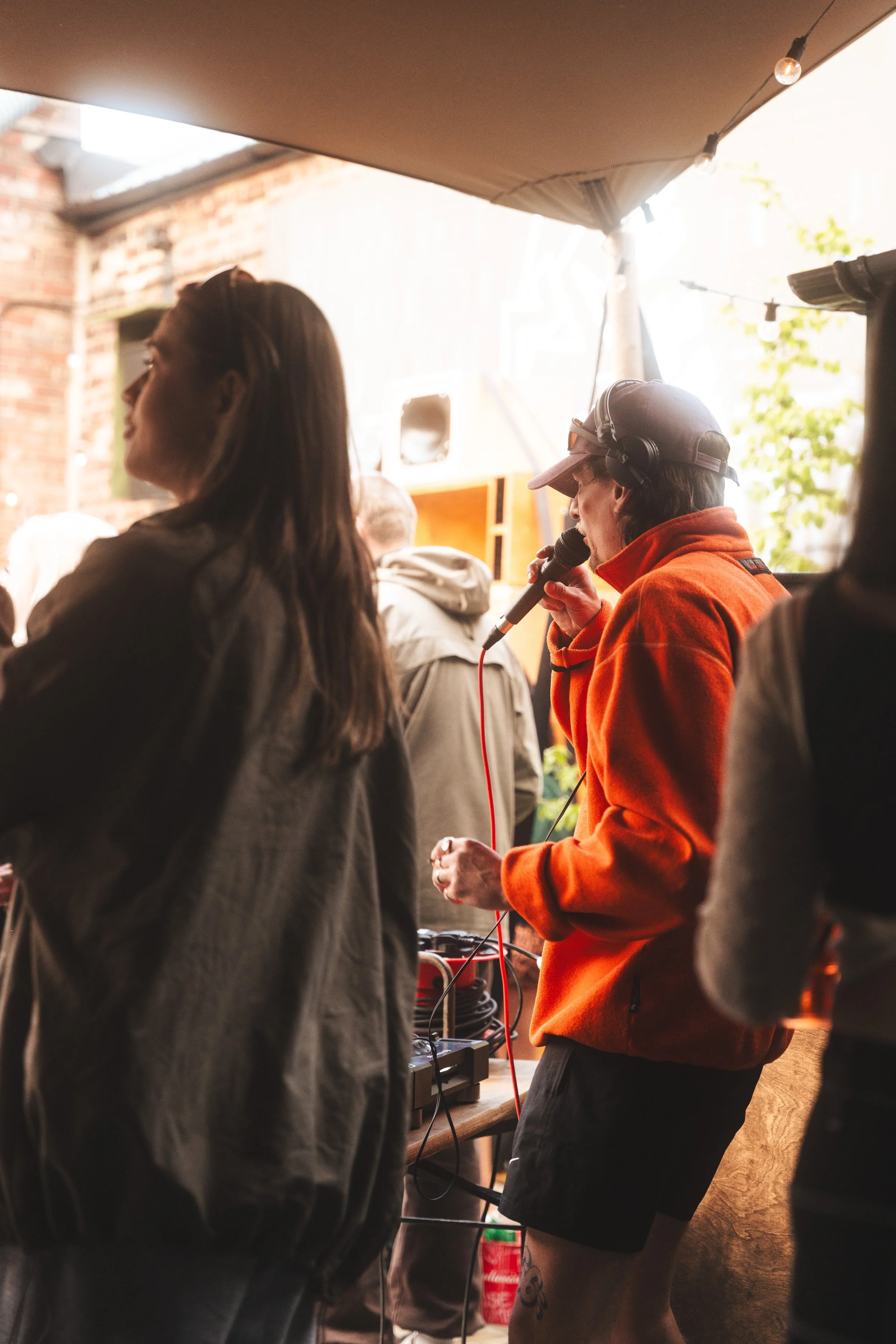 A woman in a red jacket and cap holding a microphone, performing or speaking at an outdoor event, with people standing nearby.