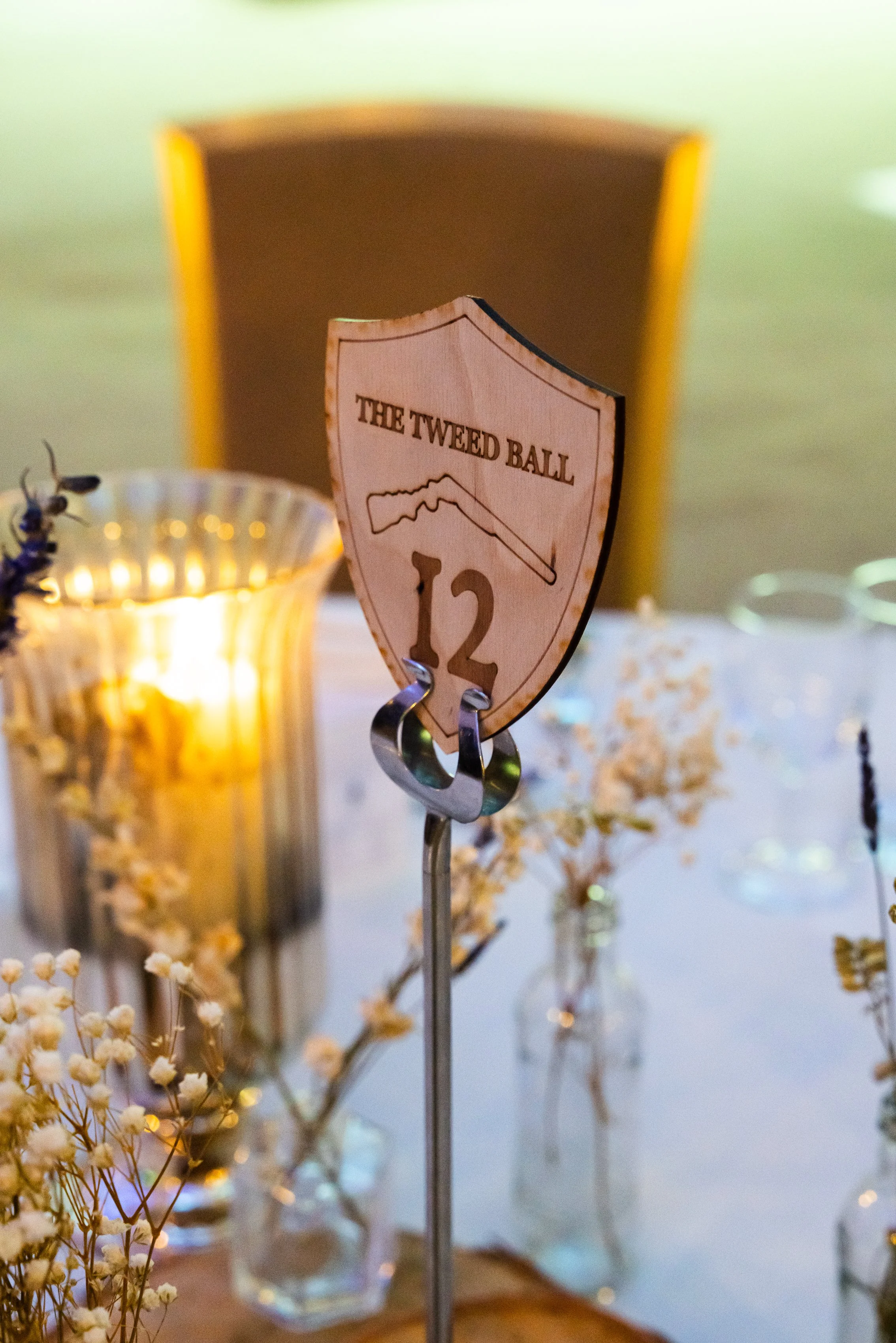 Table number sign with the text "The Tweed Ball," a mountain graphic, and the number 12, set on a table with a gold candle holder, small white flowers, and a blurred background.