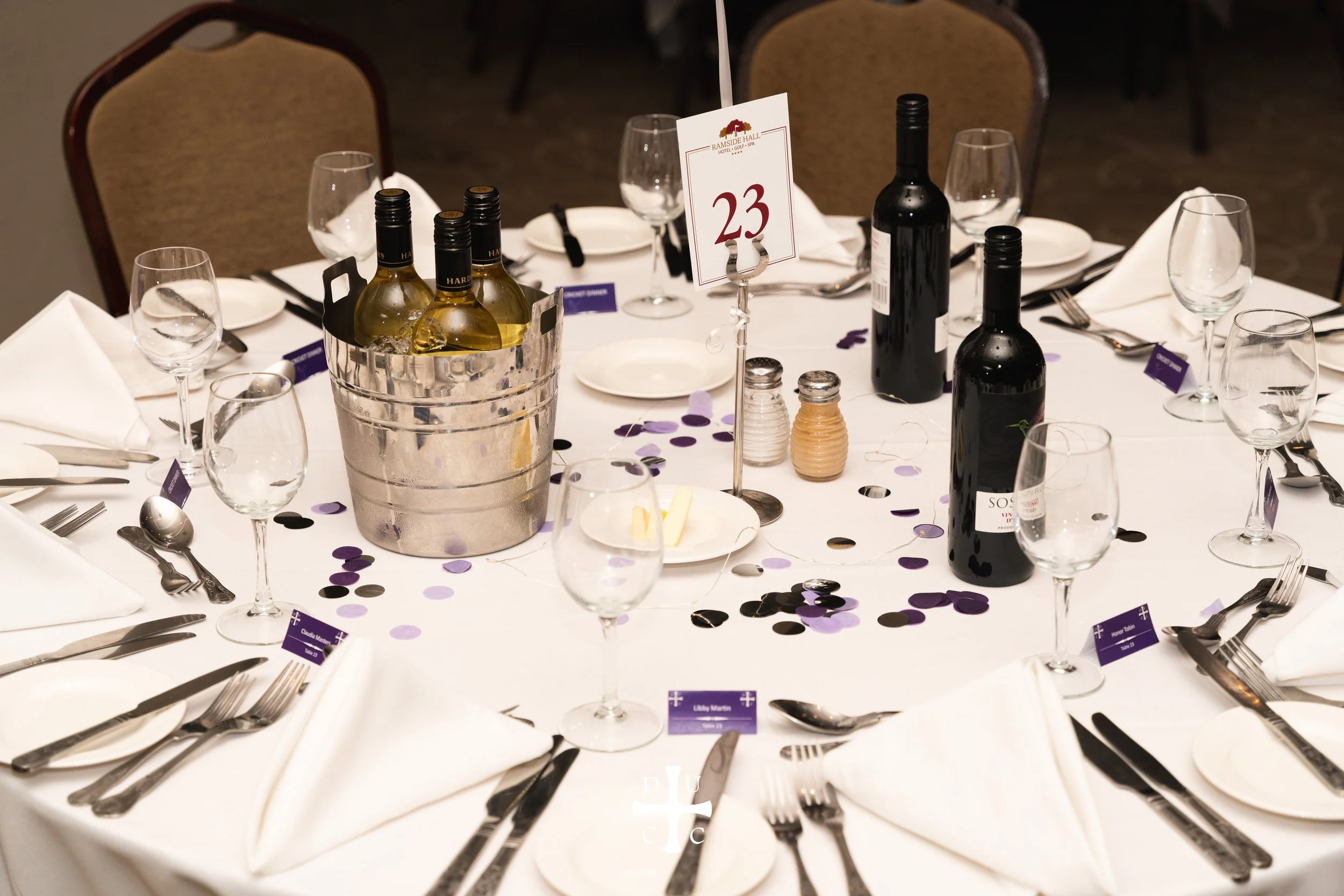 Round banquet table set with white tablecloth, white napkins, silverware, multiple wine glasses, and a centerpiece with two bottles of wine, a bucket with more wine bottles, salt and pepper shakers, and a table number card reading 23, surrounded by b