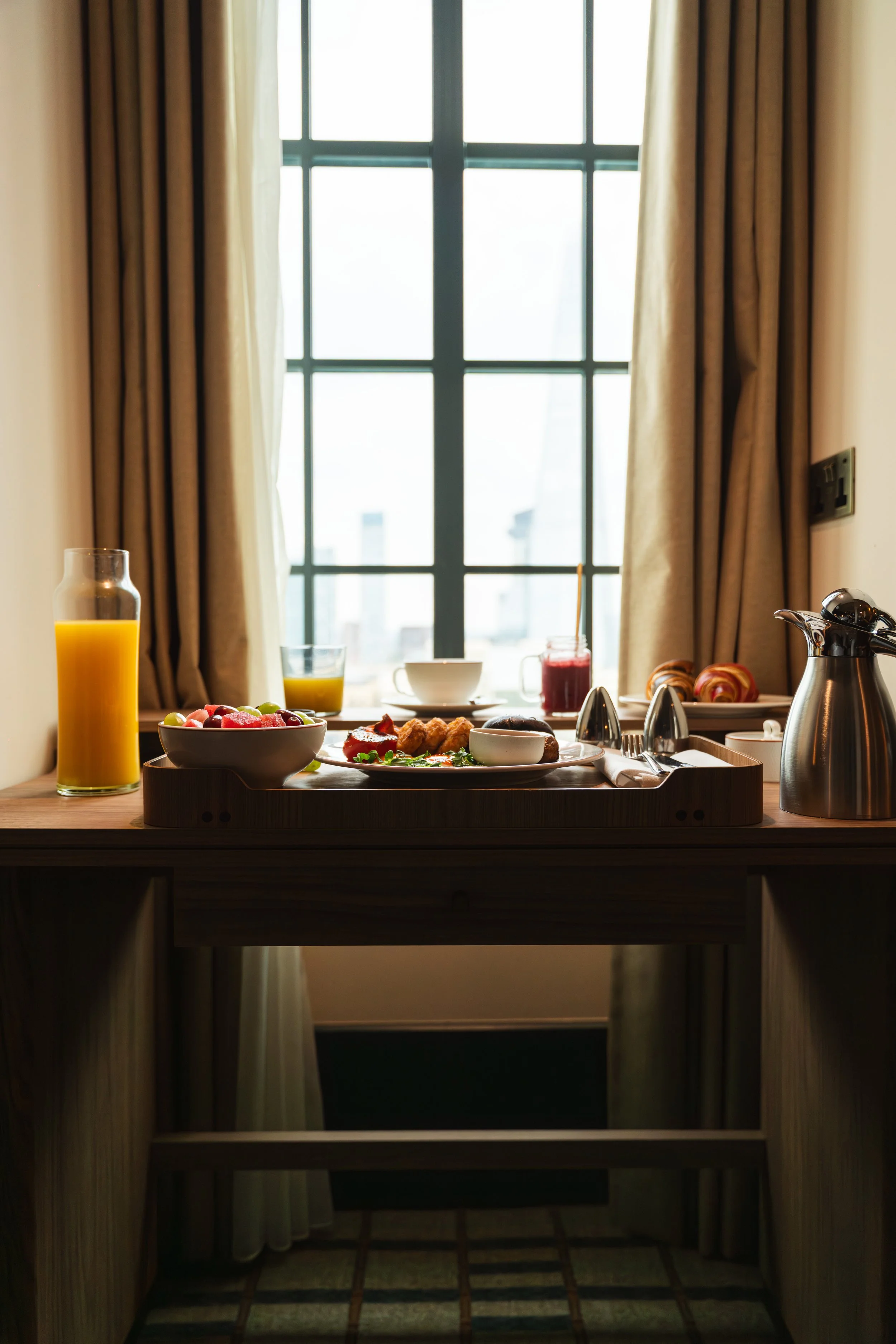 A breakfast table set by a window, with orange juice, coffee, fruit, pastries, and breakfast foods, all illuminated by natural light coming through the window.