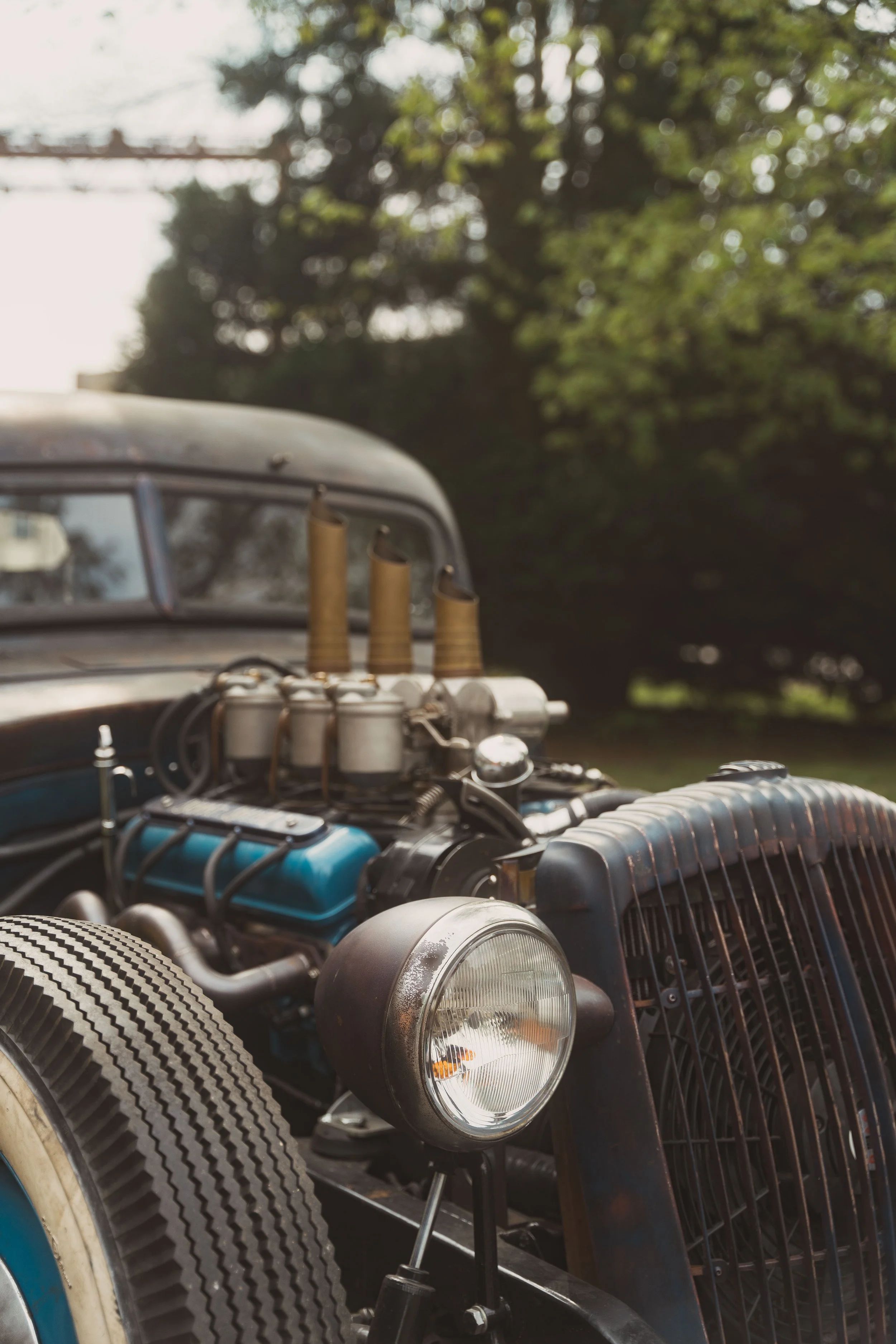 Close-up of a vintage car's front, featuring a round headlight, engine components, and a rusted metal grille, with an old tire and lush green trees in the background.