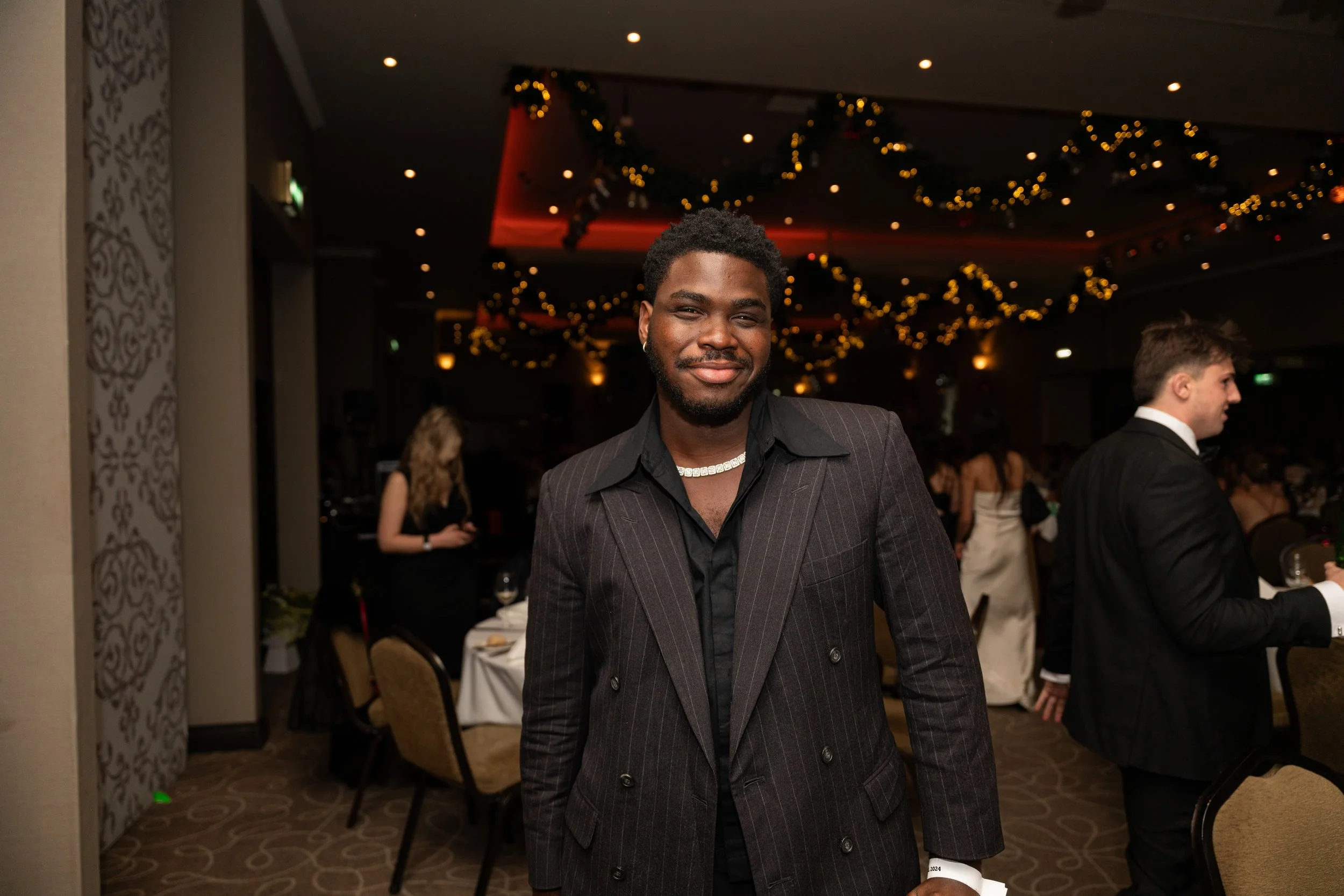 A smiling Black man in a dark suit with thin pinstripes and a black shirt stands in a decorated banquet hall, at a formal event with other guests in elegant attire, festive string lights, and holiday decorations.