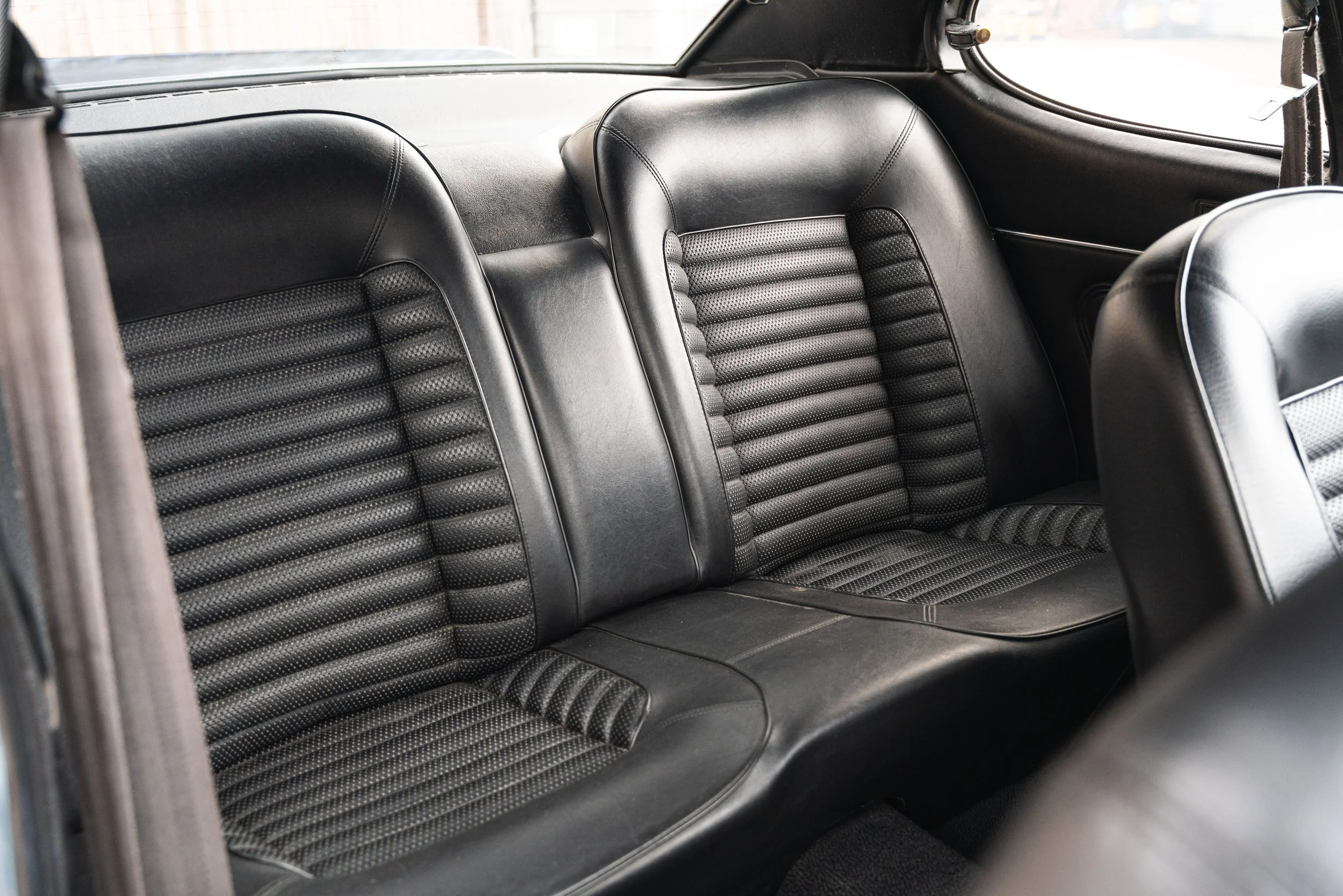 Rear black leather car seats with ribbed patterns inside a vehicle, with visible side panels and window.