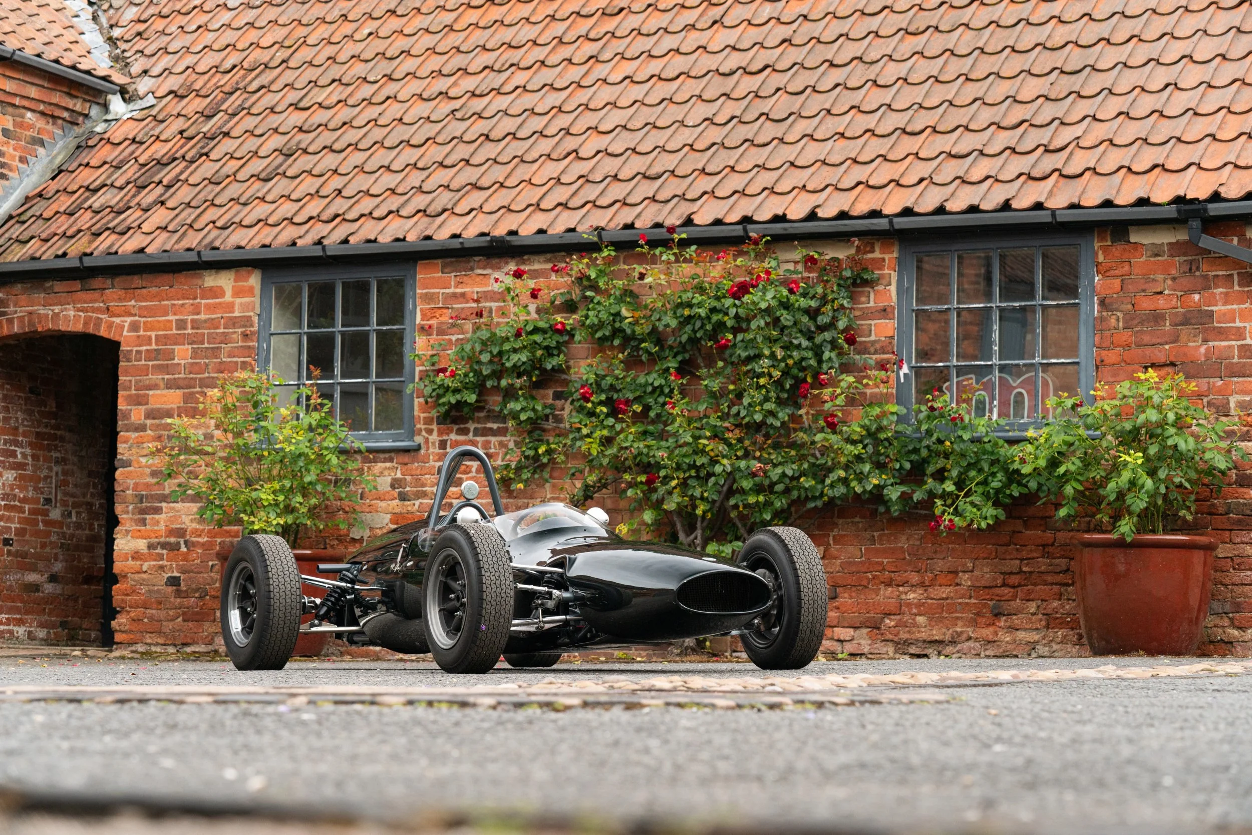 A vintage black race car parked in front of a brick house with flower pots and blooming rose bushes.