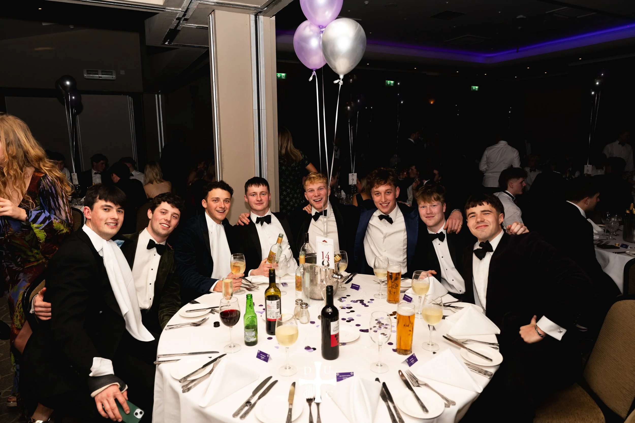 Group of young men in tuxedos sitting around a round banquet table at a formal event, smiling and holding drinks, with balloons and decorations in the background.