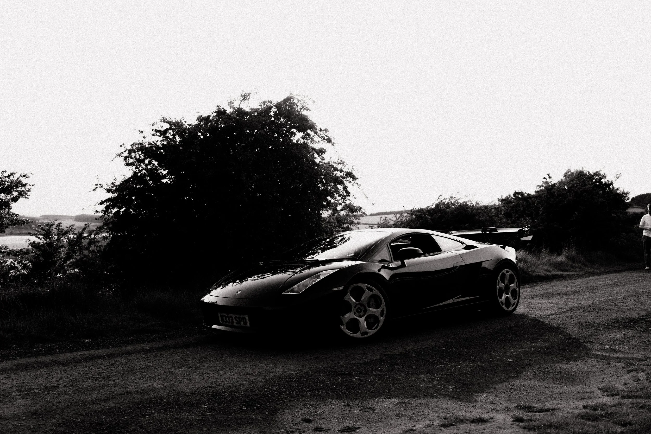A black sports car with a rear spoiler parked on a dirt road near bushes and trees, with a person standing in the background on the right side.