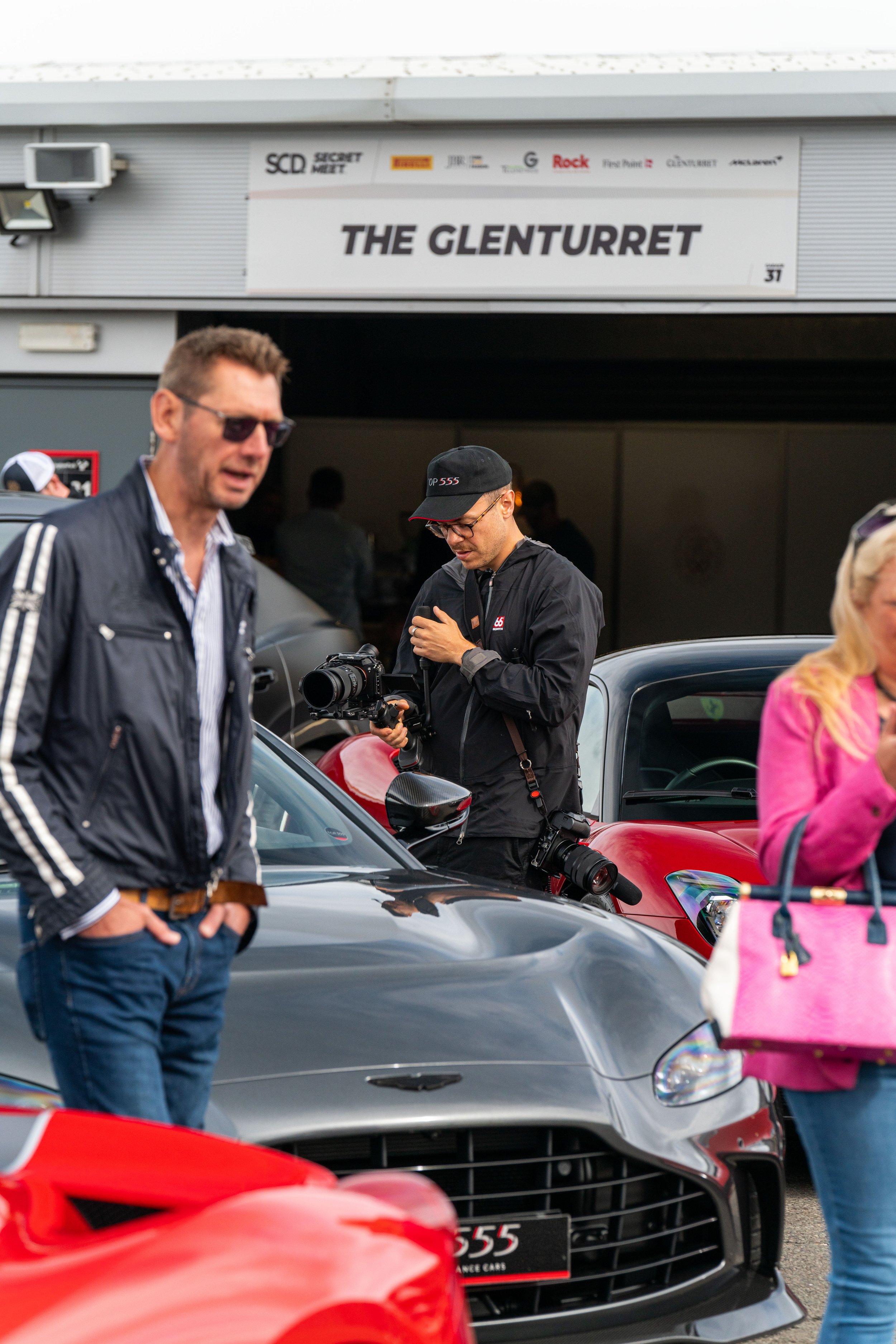 People at a car event with a sign that reads 'The Glenturret' in the background, including a man in a black jacket and cap holding a professional camera, and others around sports cars.