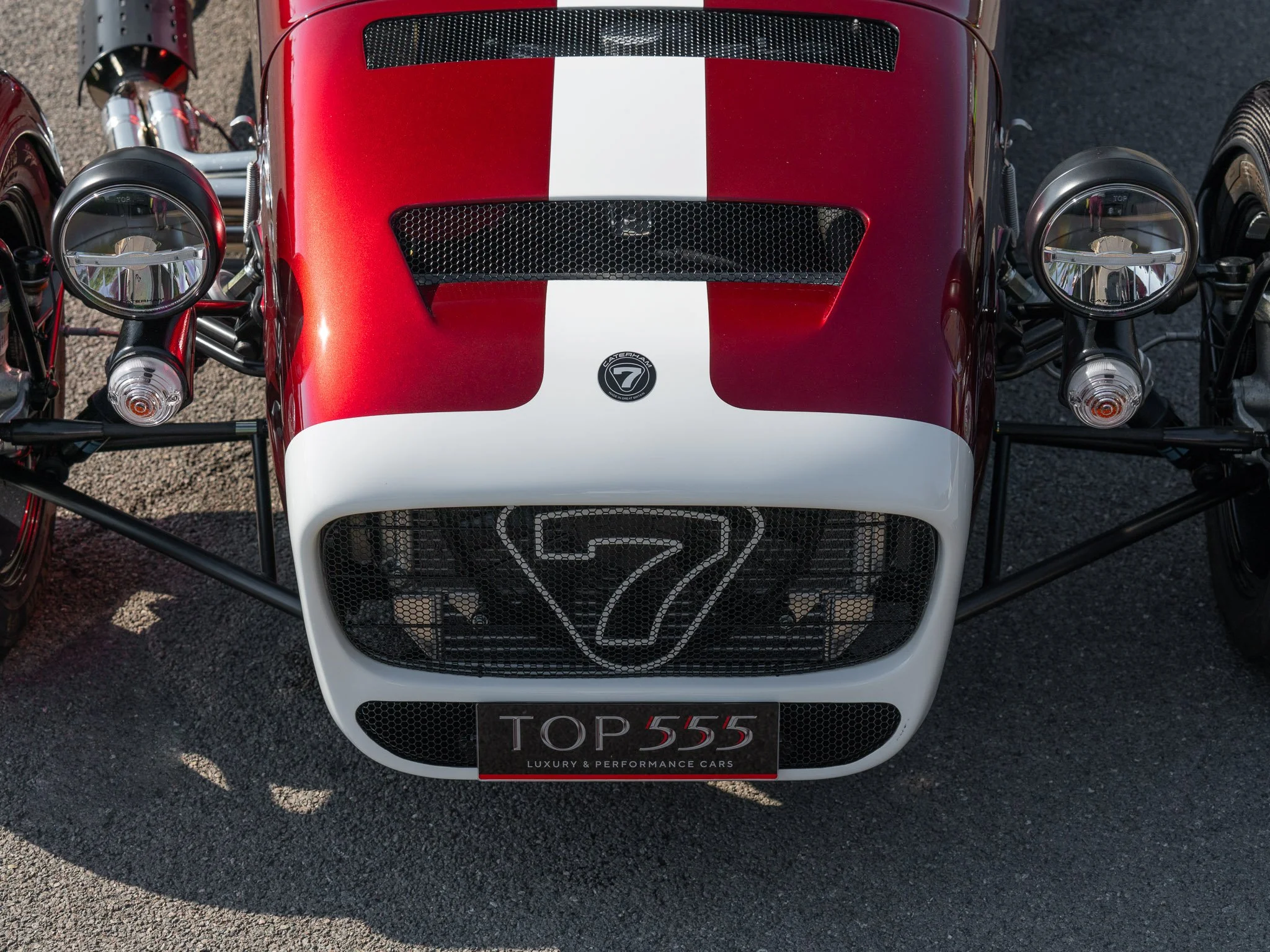 Front view of a vintage race car with a red and white body, featuring dual round headlights, a large grille with a number 7, and a license plate that reads 'TOP 555'.